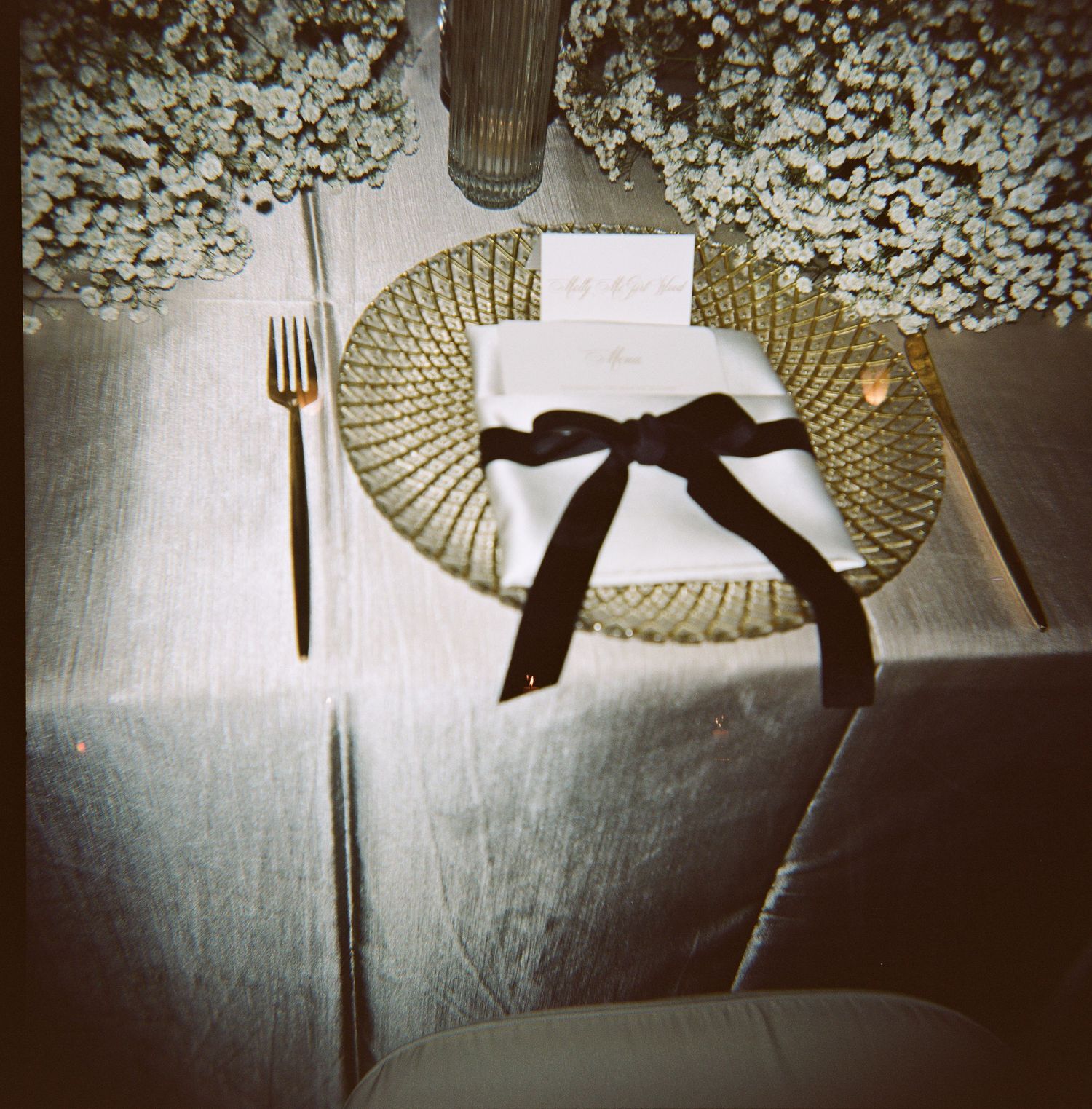 Elegant wedding table setting with a gold charger plate, white napkin tied with black velvet ribbon, place card, and baby's breath florals.