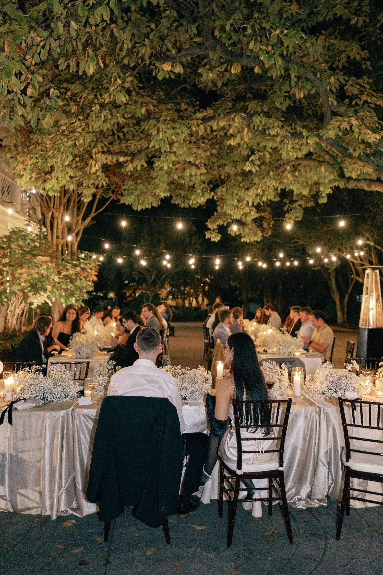 Wedding guests dine at long elegantly set tables outdoors at night under a leafy tree canopy and warm glowing string lights.
