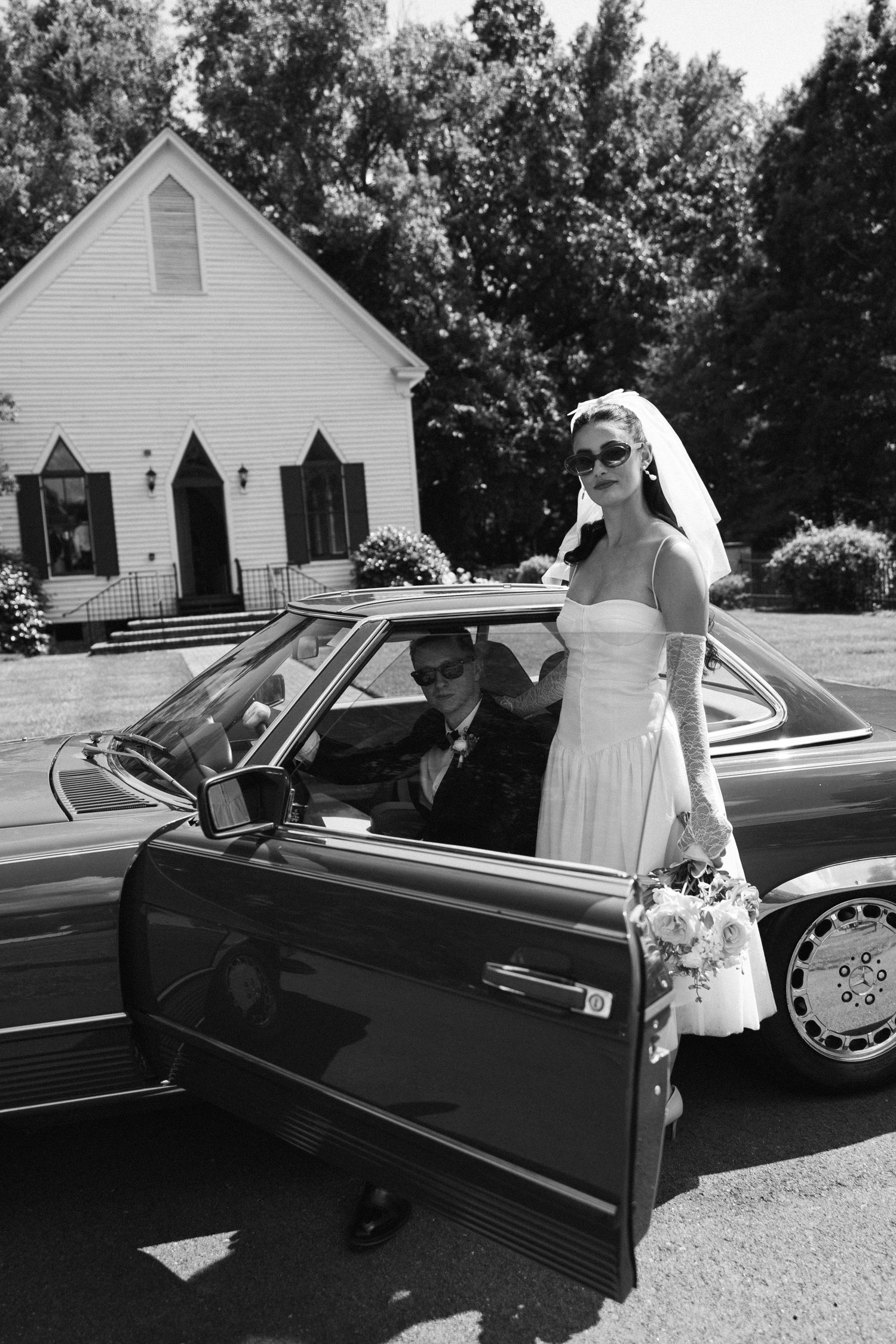 Black and white wedding photo of a bride in a veil and sunglasses beside a vintage car with the groom inside, in front of a country church.