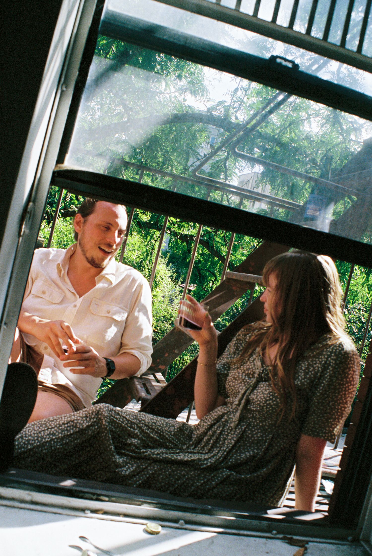 Two people relaxing and laughing together on a covered porch with lush greenery visible through windows.