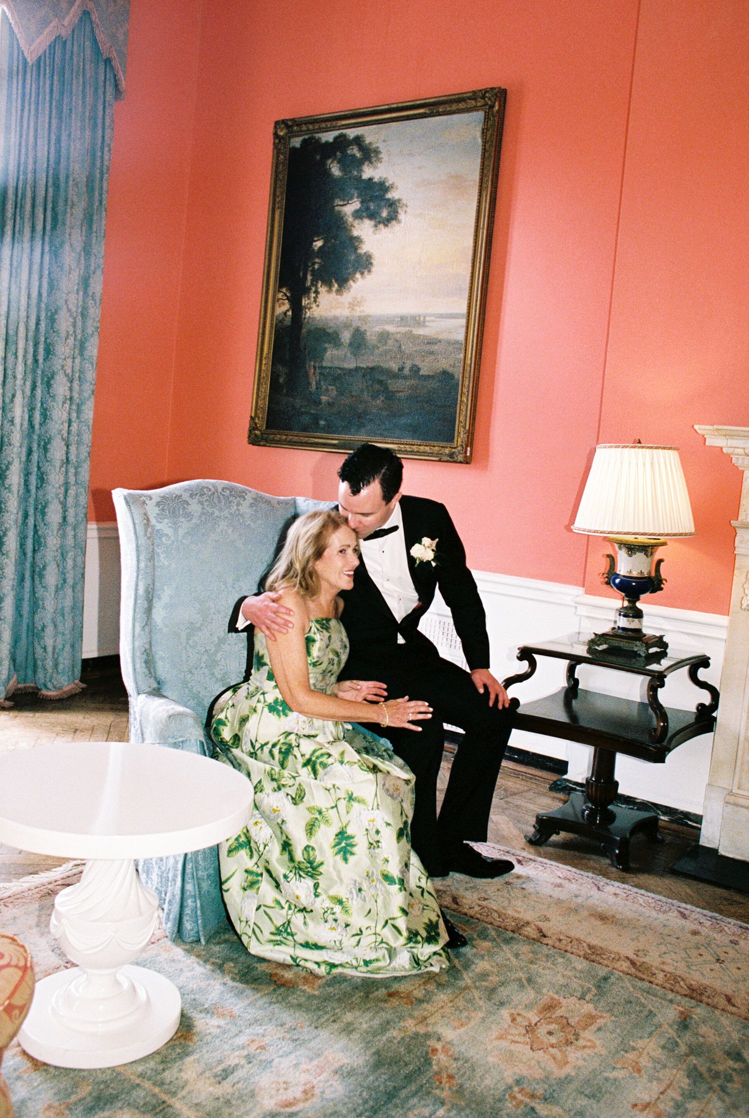 Bride in floral gown and groom in tuxedo share intimate moment in elegant coral room with classic painting.