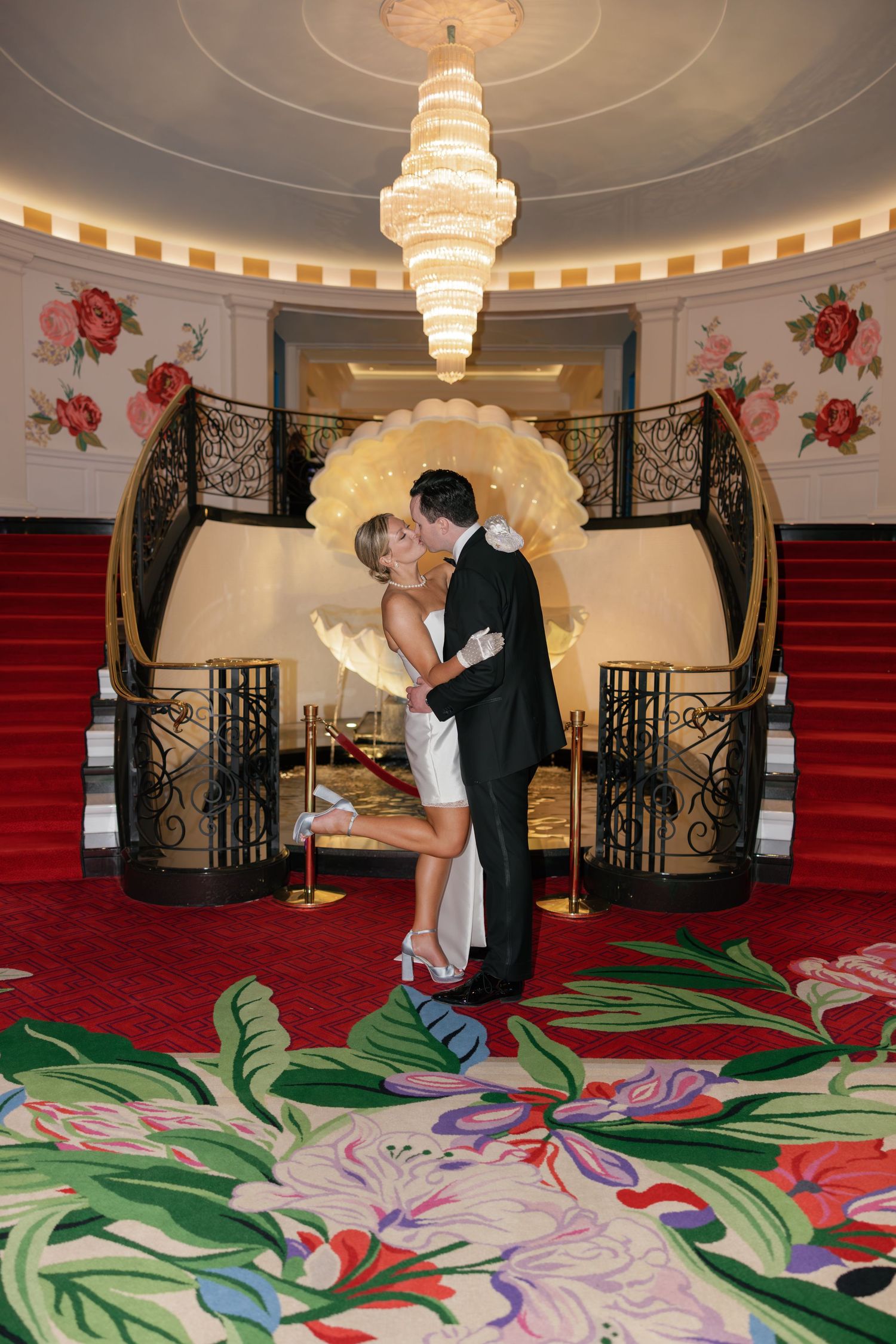 Couple sharing a romantic kiss in an elegant ballroom with grand staircase, chandelier, and floral carpet.