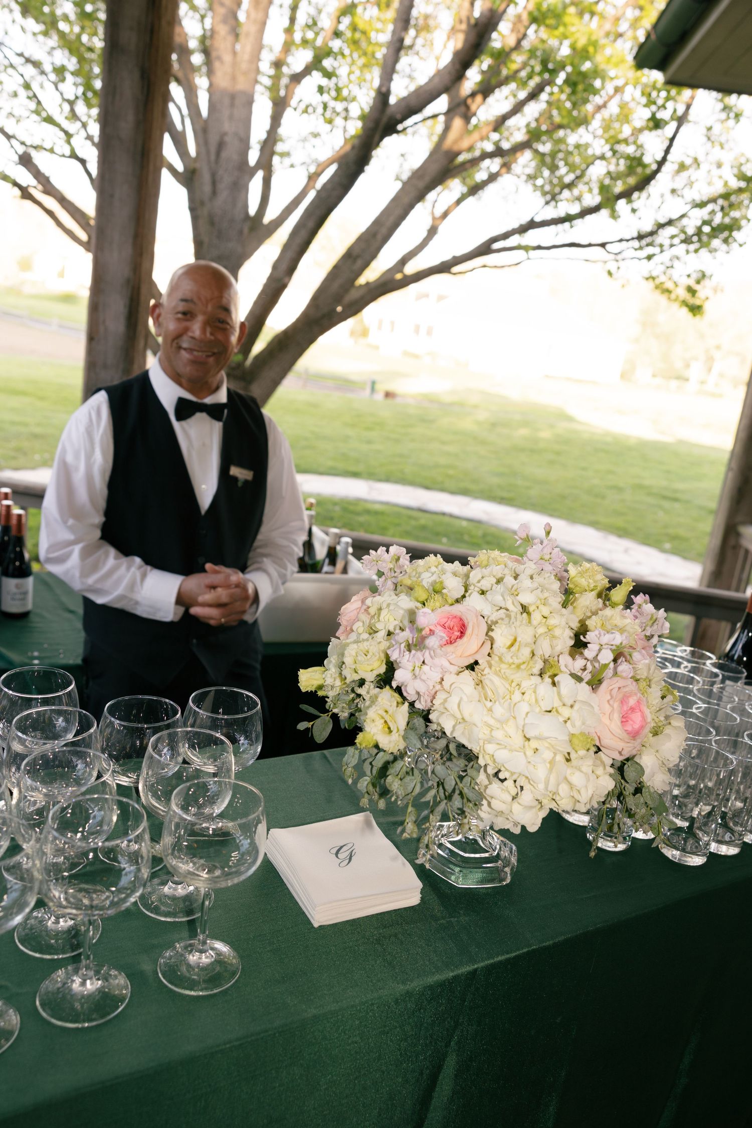 A smiling bartender in a bow tie stands behind a green-draped table with crystal glasses and floral arrangement.