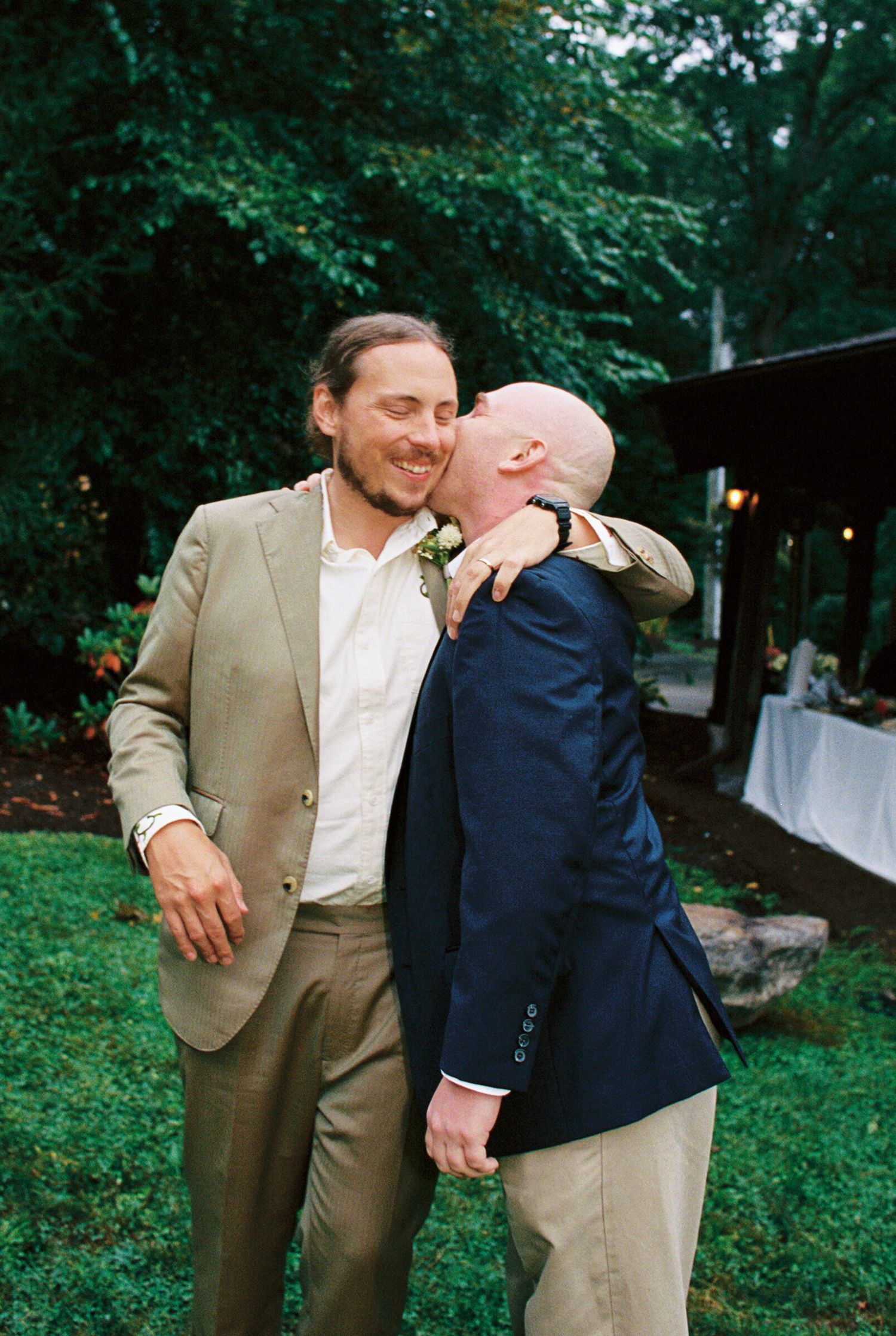Two men share a joyful moment at an outdoor wedding celebration with green trees and white decor in the background.