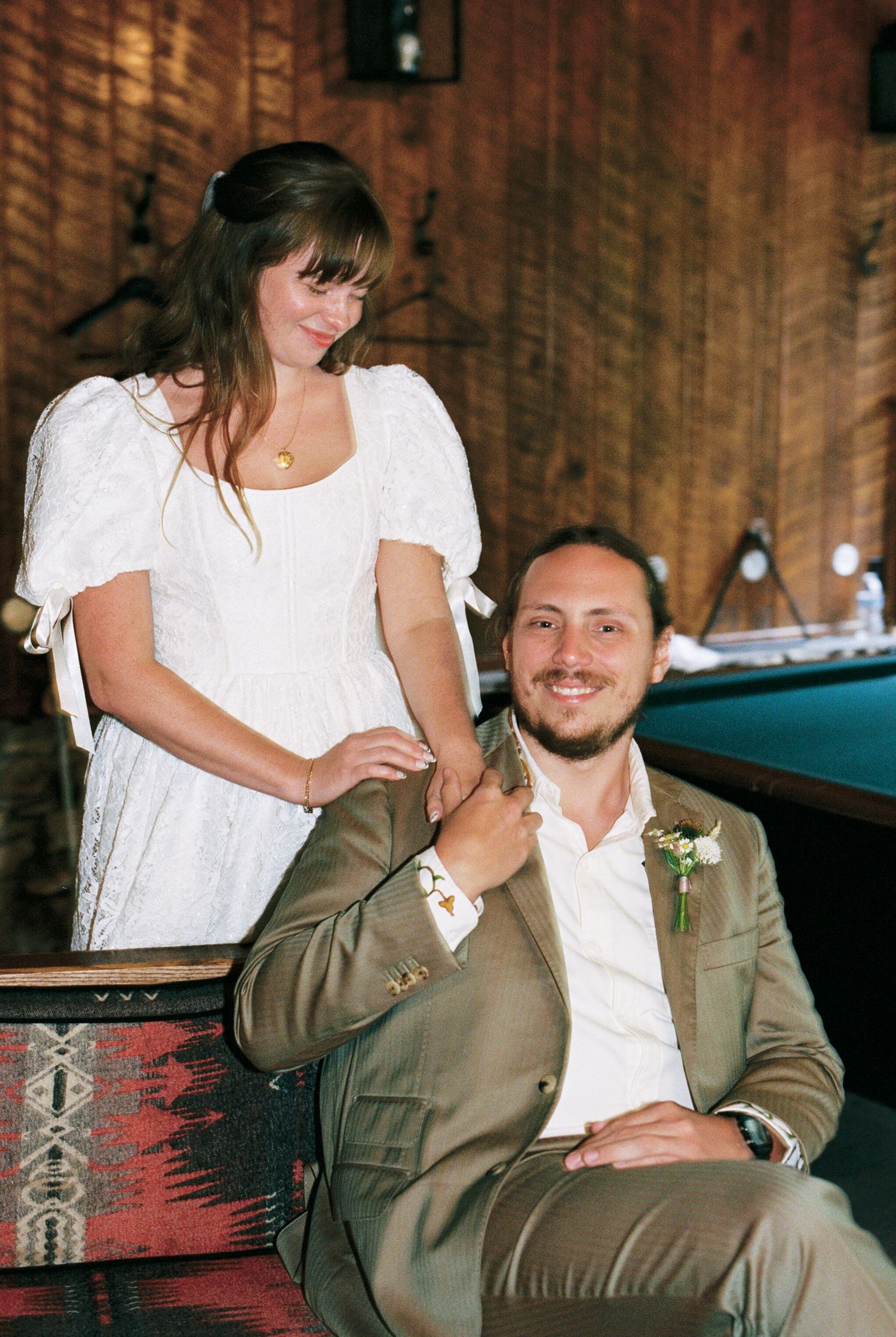 A couple sits together on a rustic wooden bench wearing white and olive green attire in a romantic outdoor setting.