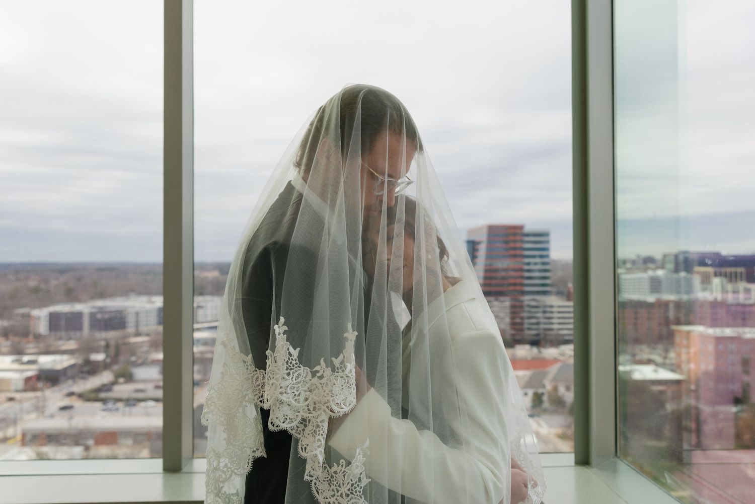Bride and groom share intimate moment under lace wedding veil by floor-to-ceiling windows with city skyline backdrop.