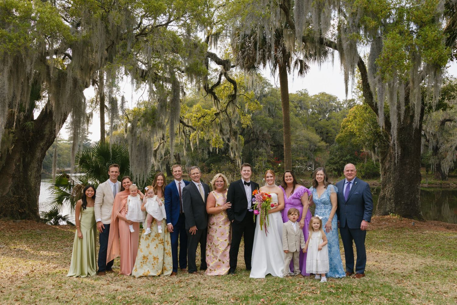 Family wedding portrait group posed outdoors under Spanish moss-draped trees near a pond, featuring bride and groom with guests.