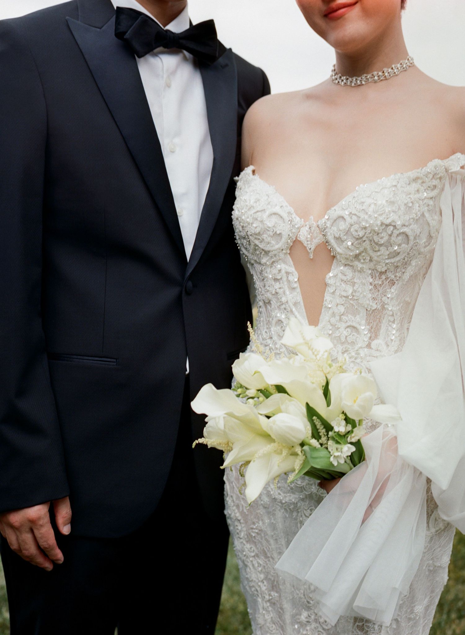 Close-up of formal wedding attire showing black tuxedo and detailed white lace gown with bouquet.