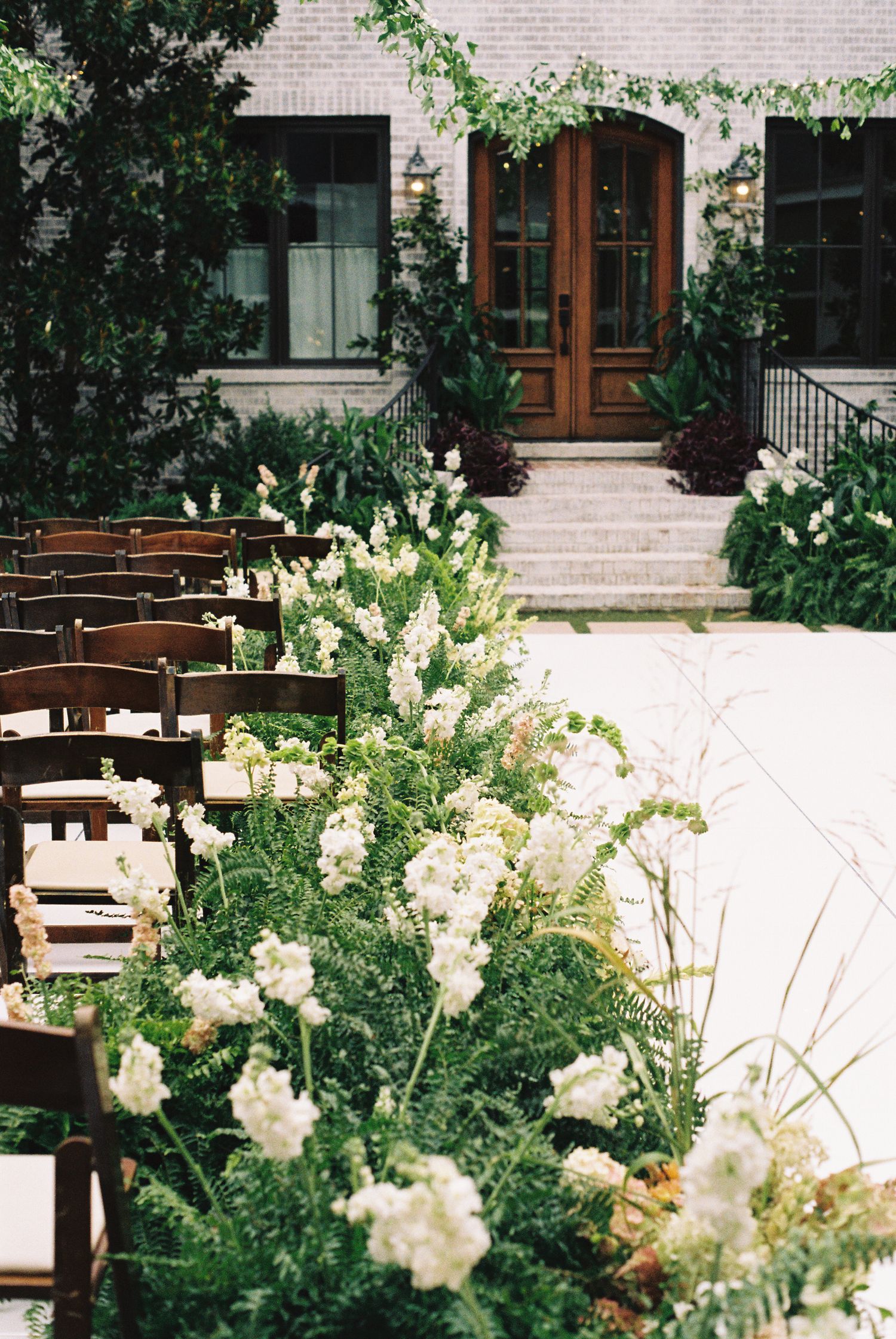 Outdoor wedding ceremony setup with white floral arrangements lining the aisle, wooden chairs, and elegant building entrance.