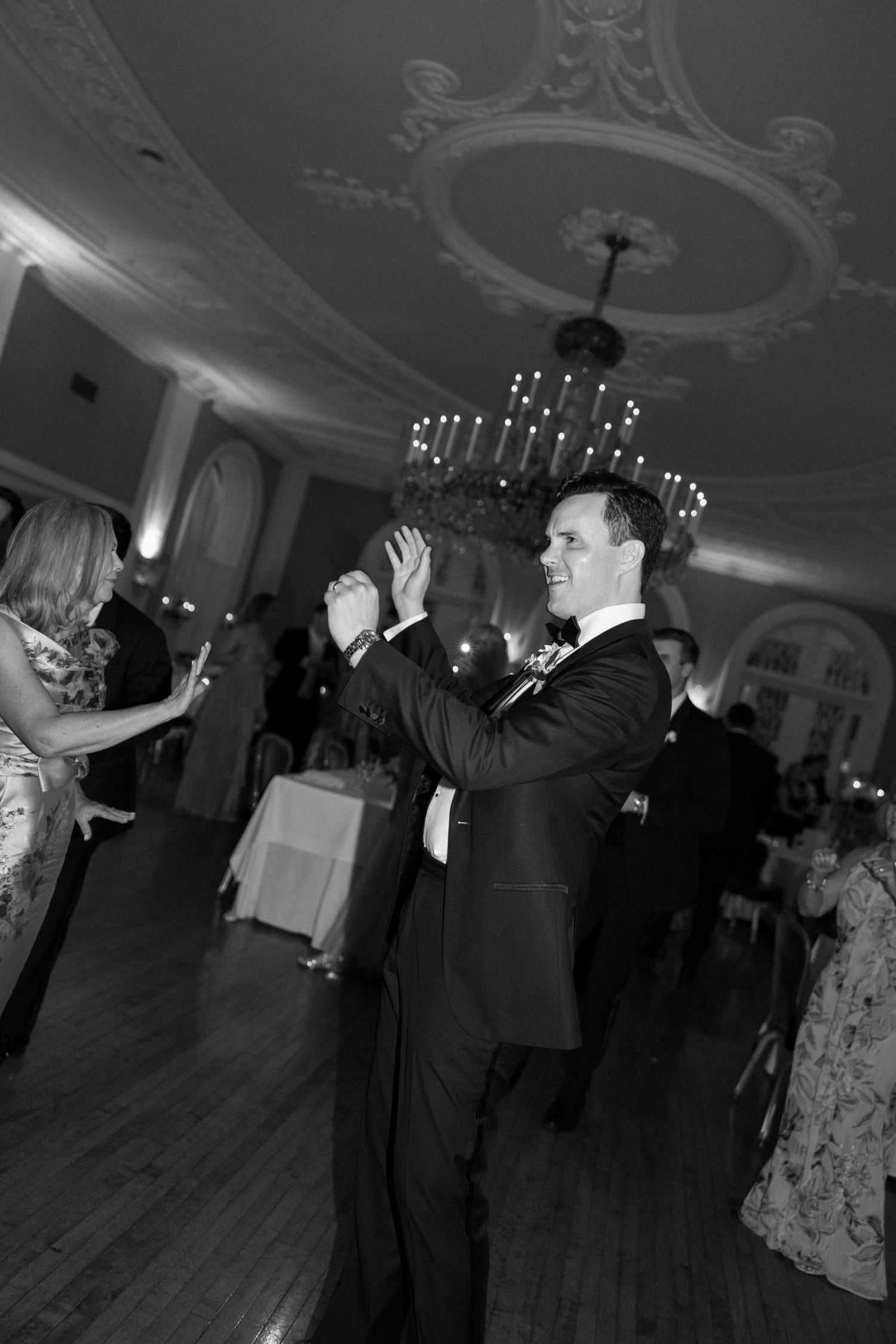 Groom smiling and dancing at elegant black and white wedding reception with ornate ceiling and chandelier.