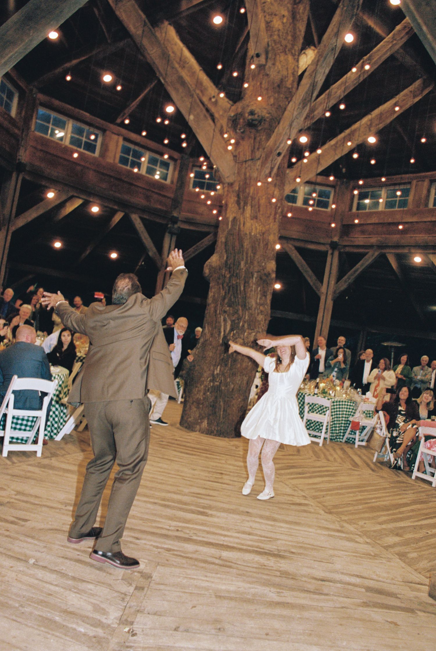 Wedding guests dance and celebrate on a rustic wooden barn floor illuminated by string lights overhead.