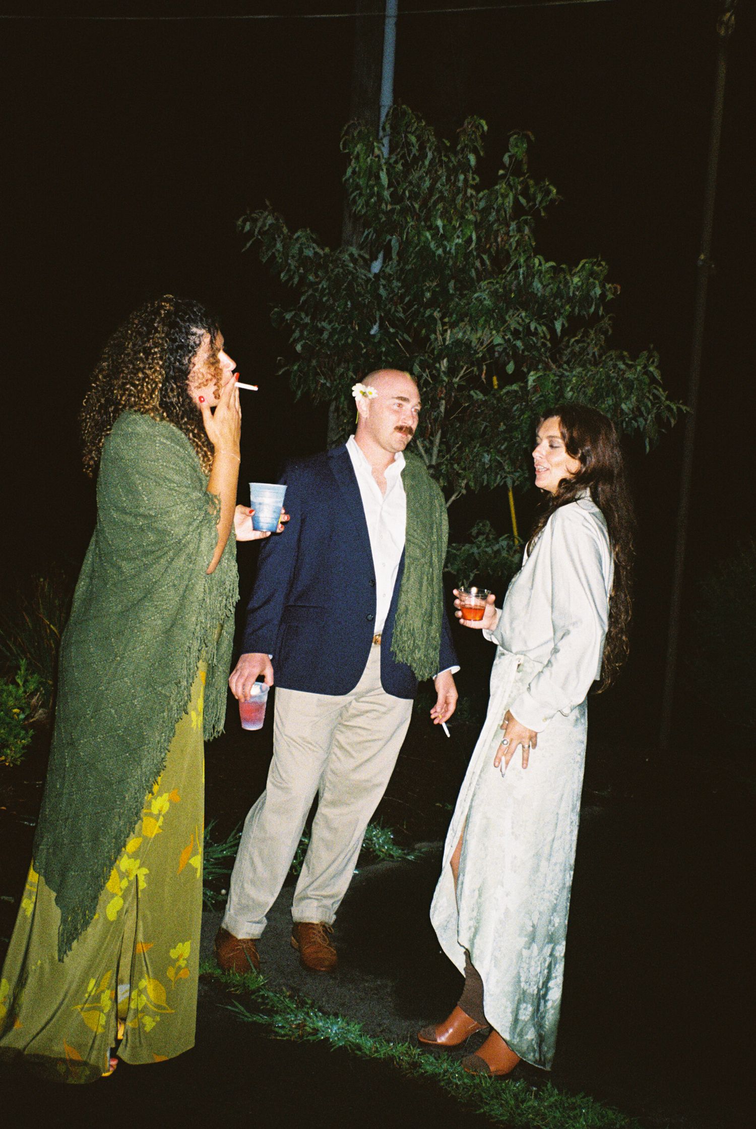 Three people having a casual conversation at a nighttime outdoor gathering, with two wearing long dresses and one in a blazer.