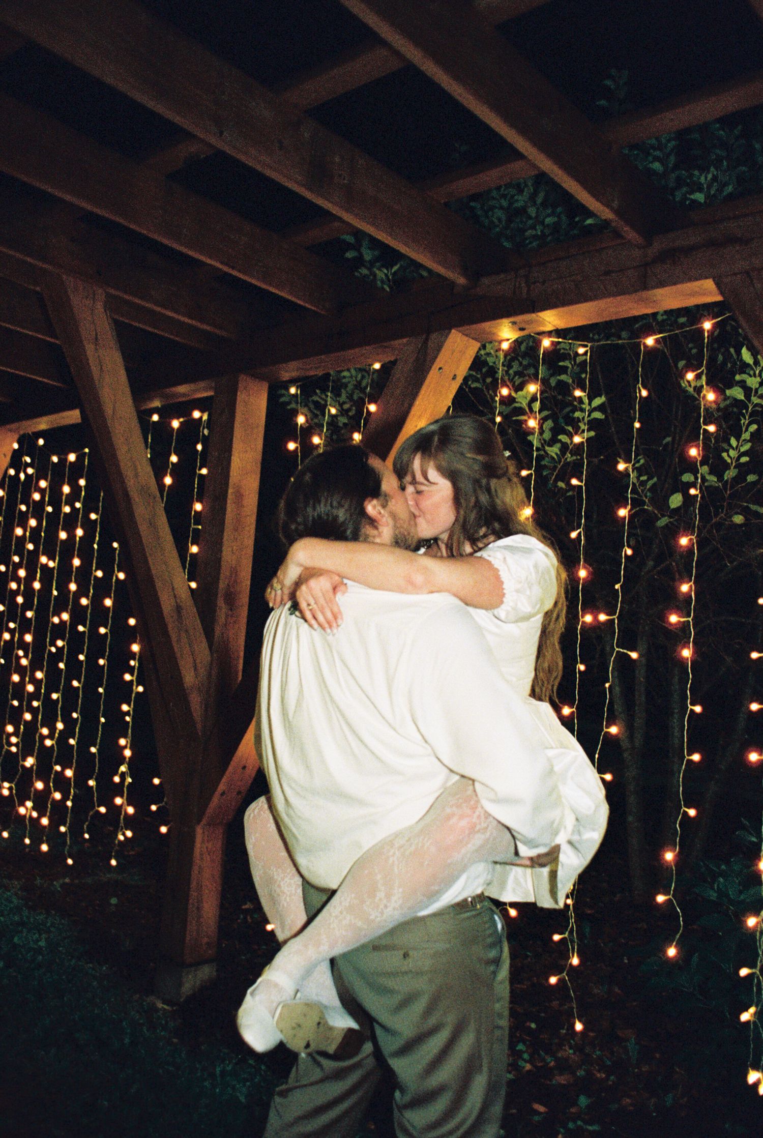 A romantic couple shares a dance and kiss under twinkling string lights in a rustic wooden gazebo at dusk.