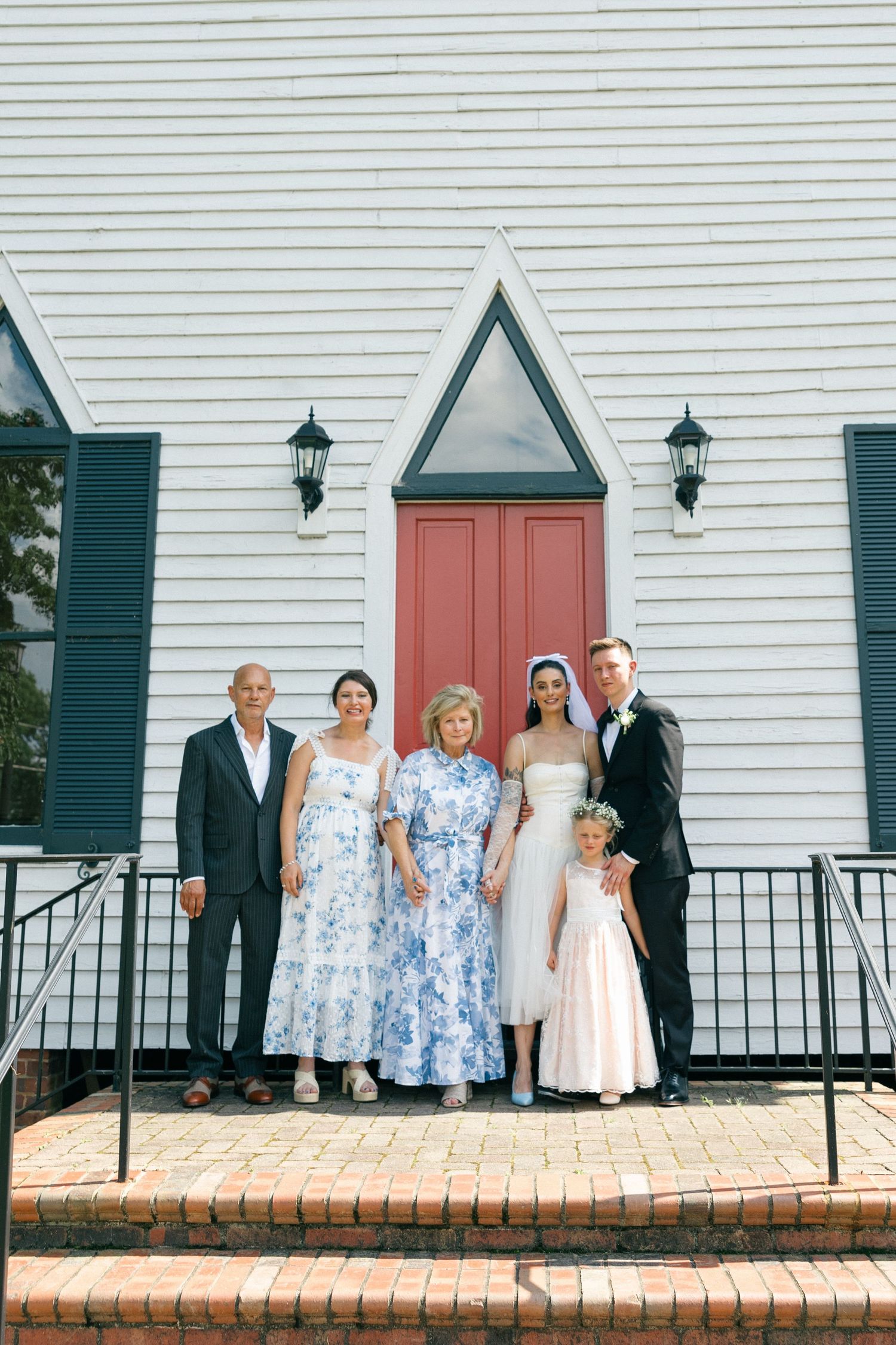 Bride and groom pose with a flower girl and family members on brick steps outside a white wooden church with red doors.