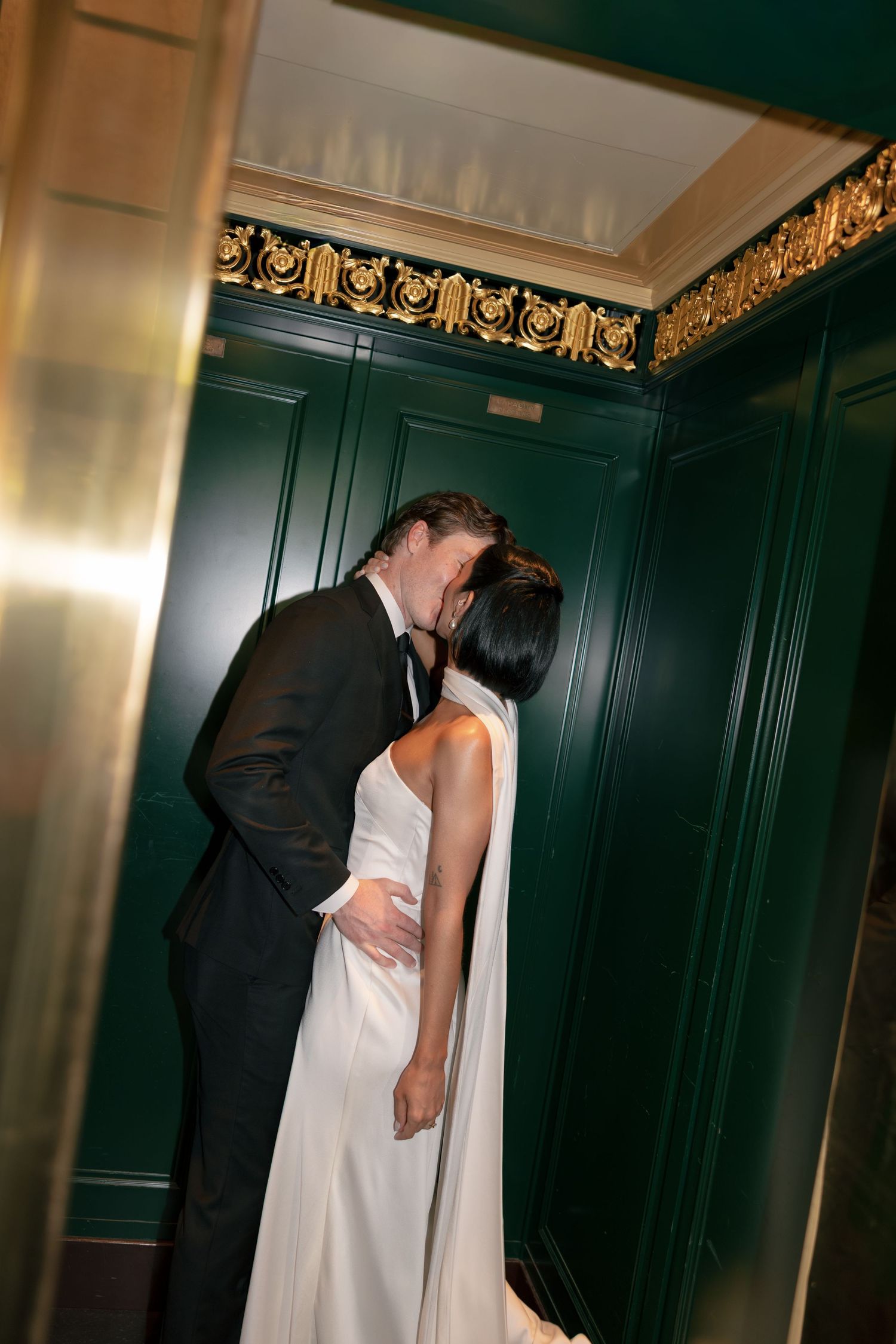 A bride in a white satin gown and veil kisses a groom in a black suit inside an elegant green paneled elevator with gold trim.
