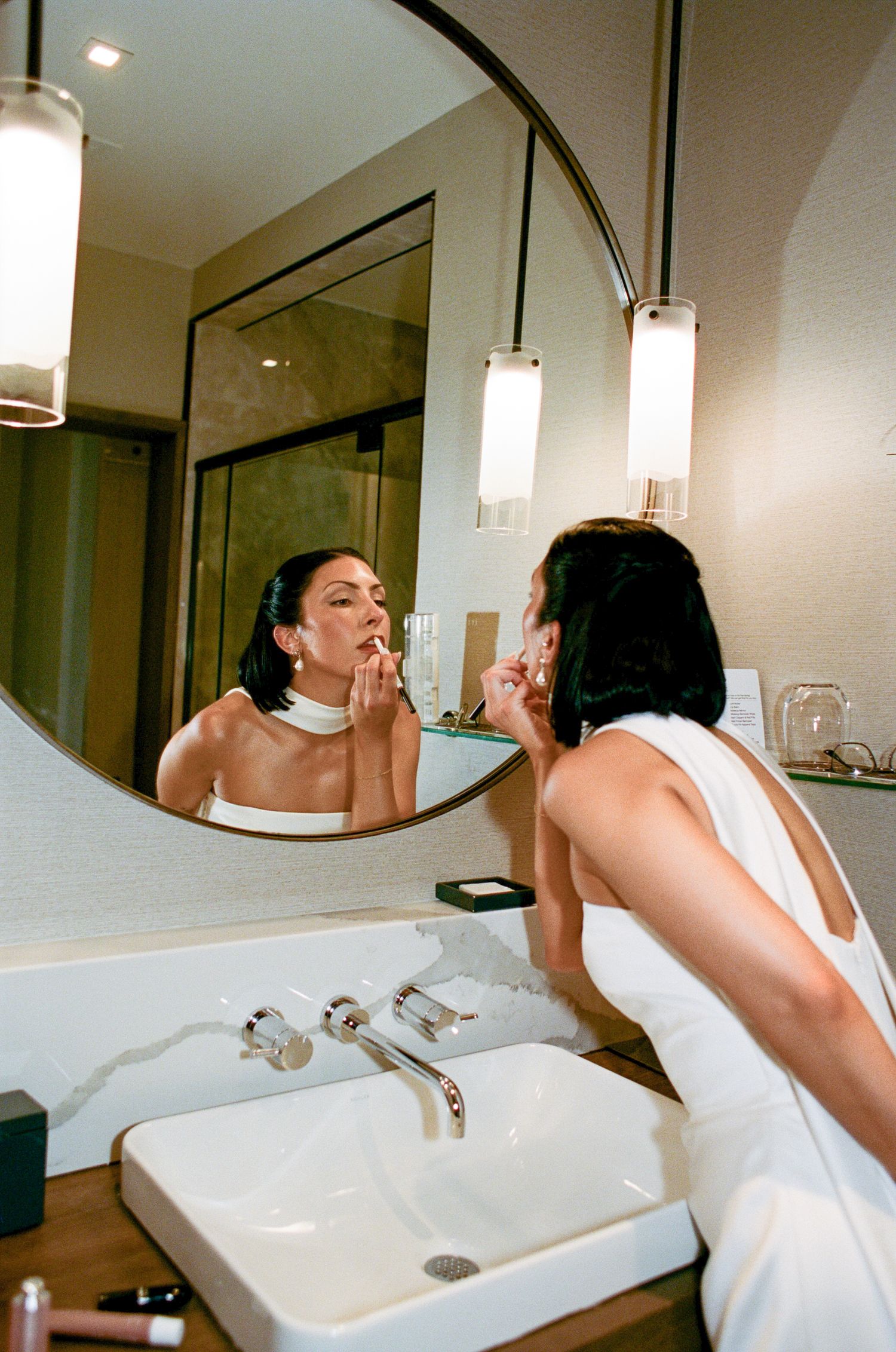 Woman in a white halter top applies lipstick while looking into a round mirror above a modern white sink with pendant lights.