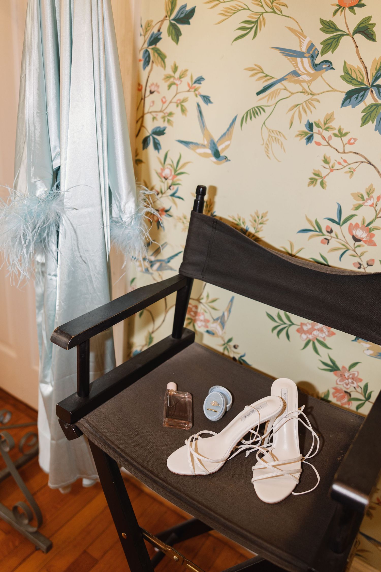 Bridal accessories including strappy heels, perfume, and a blue brooch on a black director's chair with floral wallpaper.