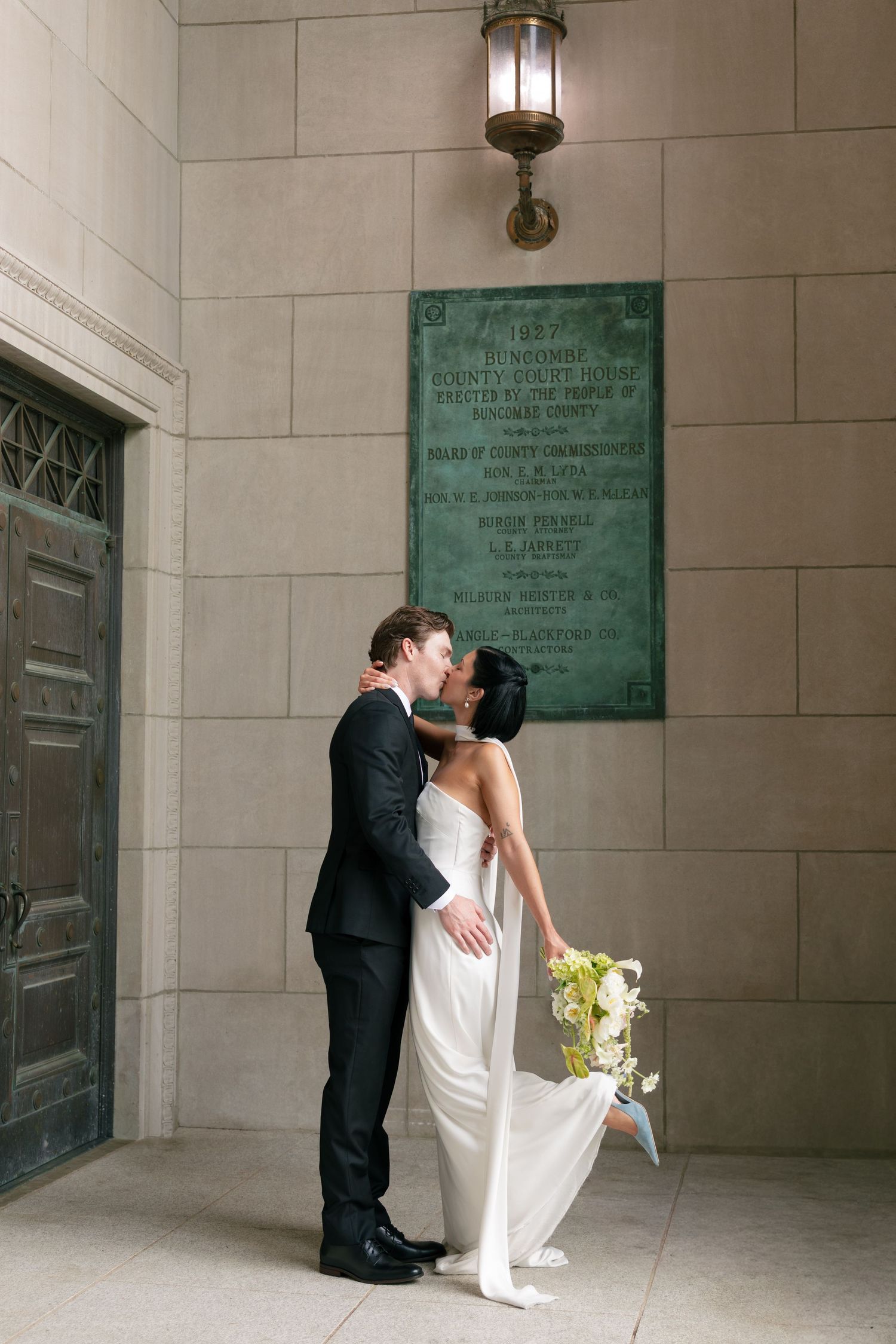 Bride and groom share a kiss outside a stone building, bride holding white flowers in an elegant white gown.
