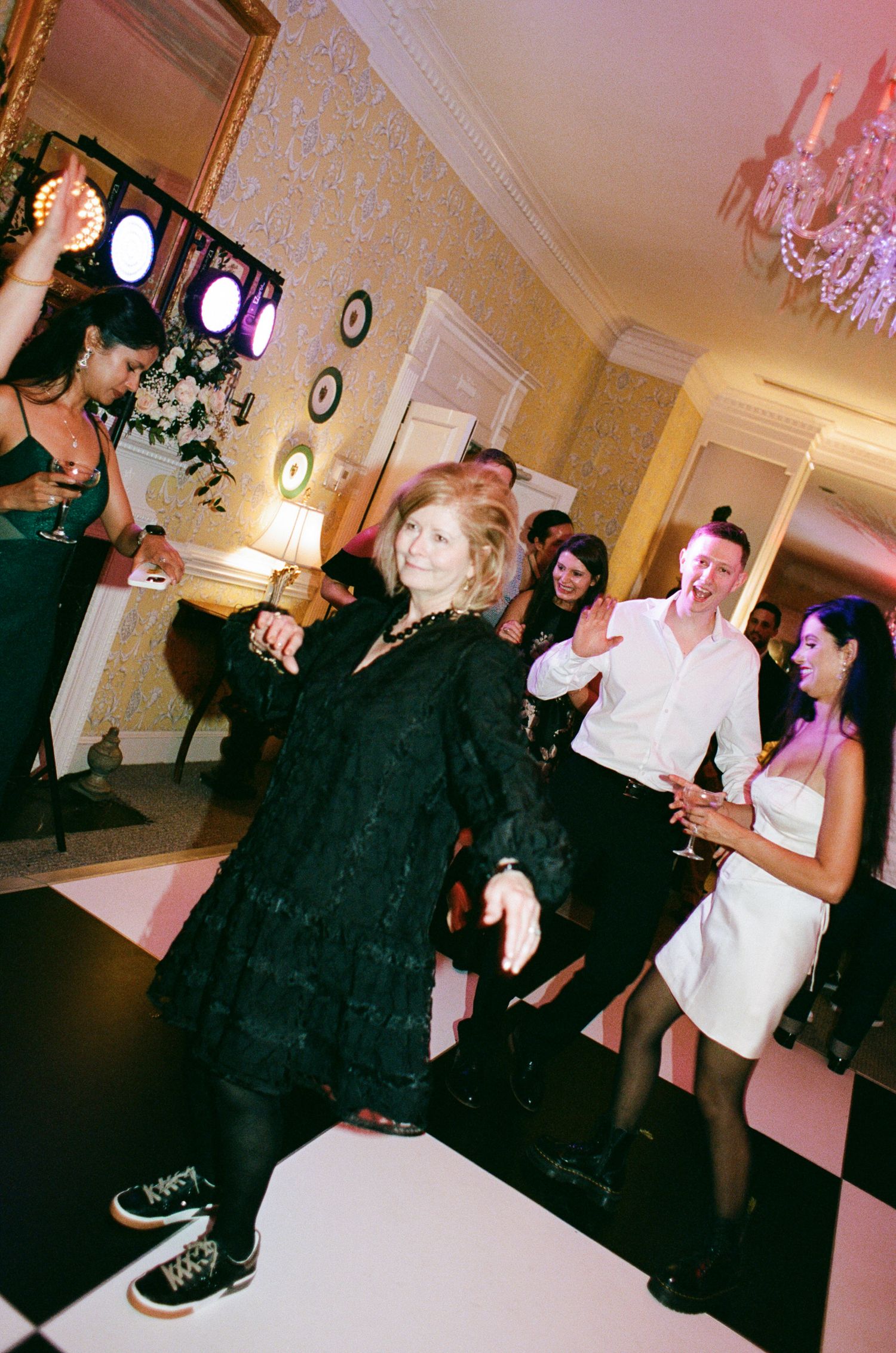 Guests dance and celebrate on a black and white checkered floor at an elegant wedding reception with a chandelier and stage lights.