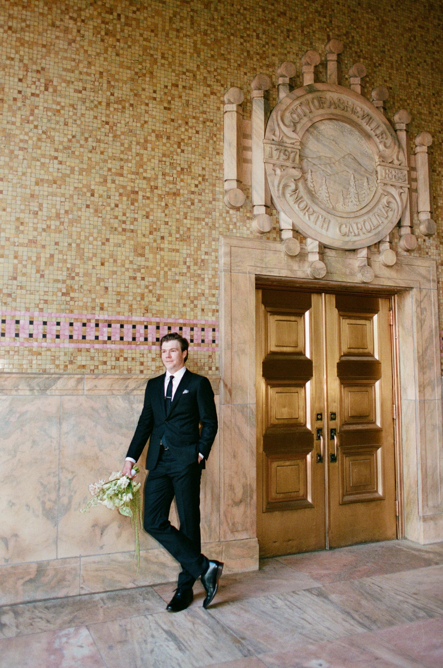 Groom in a black suit and tie holds a white bouquet while standing beside ornate wooden doors and a carved stone seal in a marble hall.