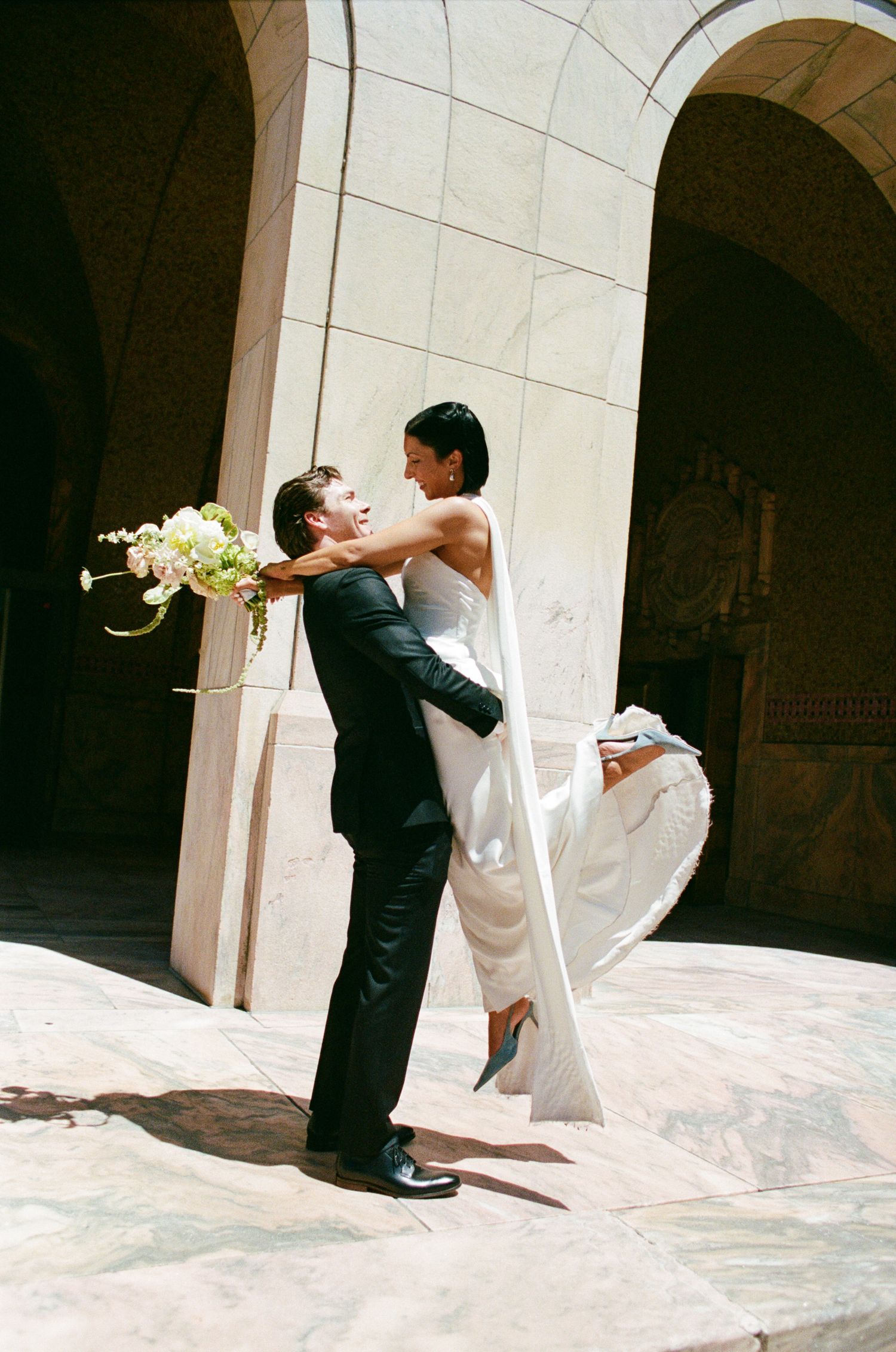 Groom in a black suit lifts a smiling bride in a white gown, holding a bouquet, beneath stone archways on a sunlit marble plaza.