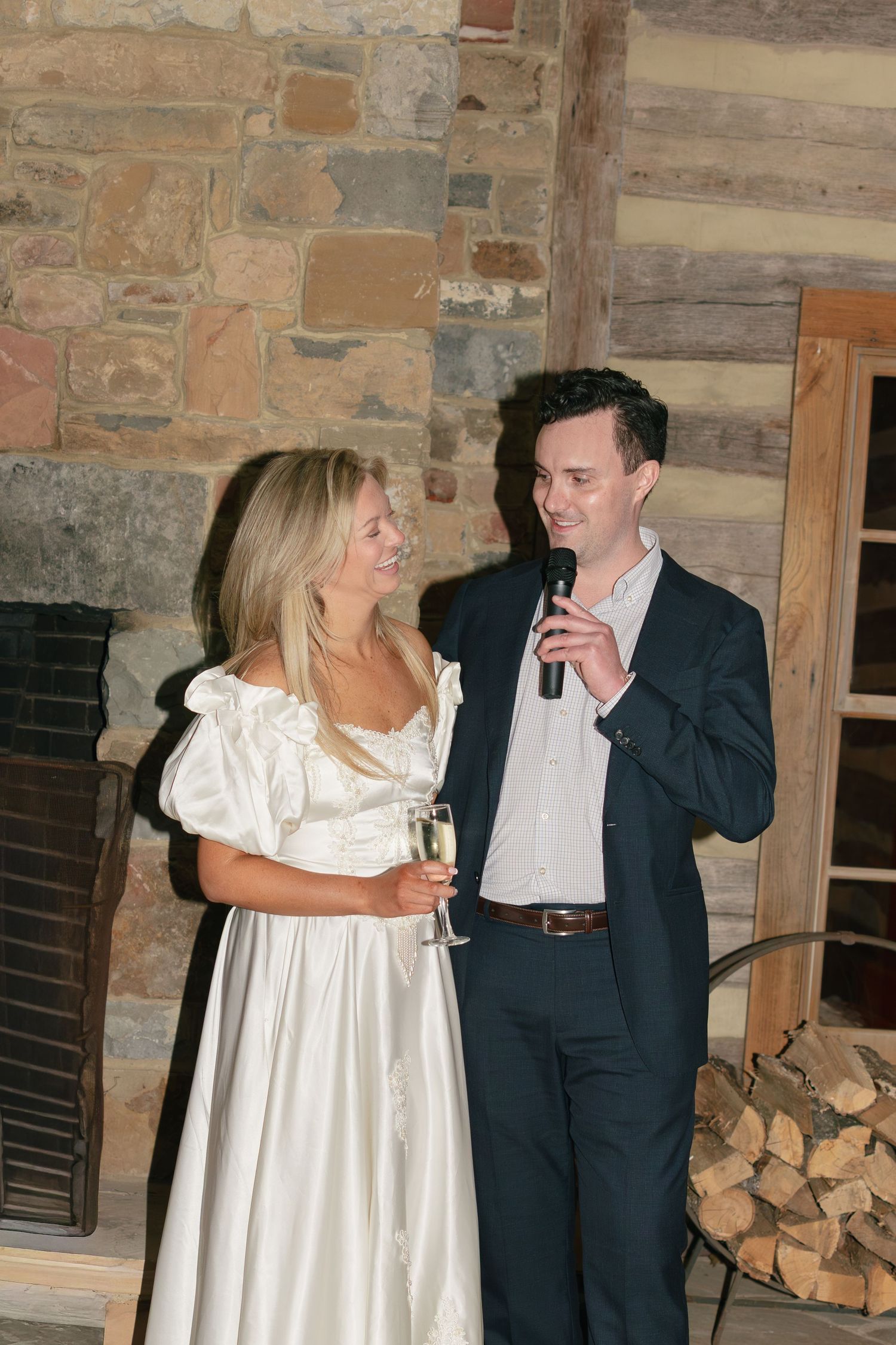 Bride in white satin gown and groom in navy suit giving champagne toast speech at rustic stone wedding venue.