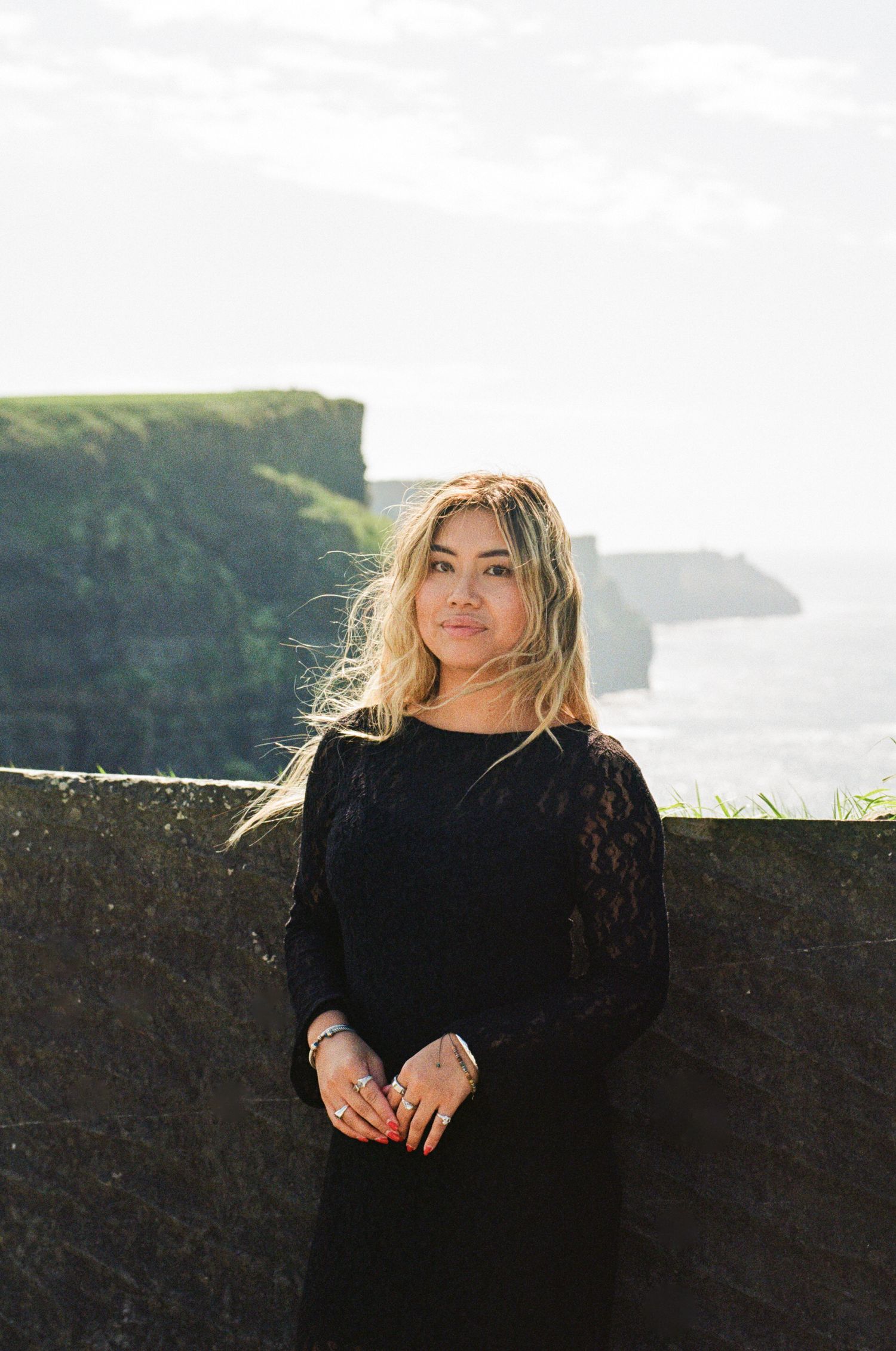 Woman in black lace dress standing at Cliffs of Moher, Ireland, with dramatic coastal cliffs in background.