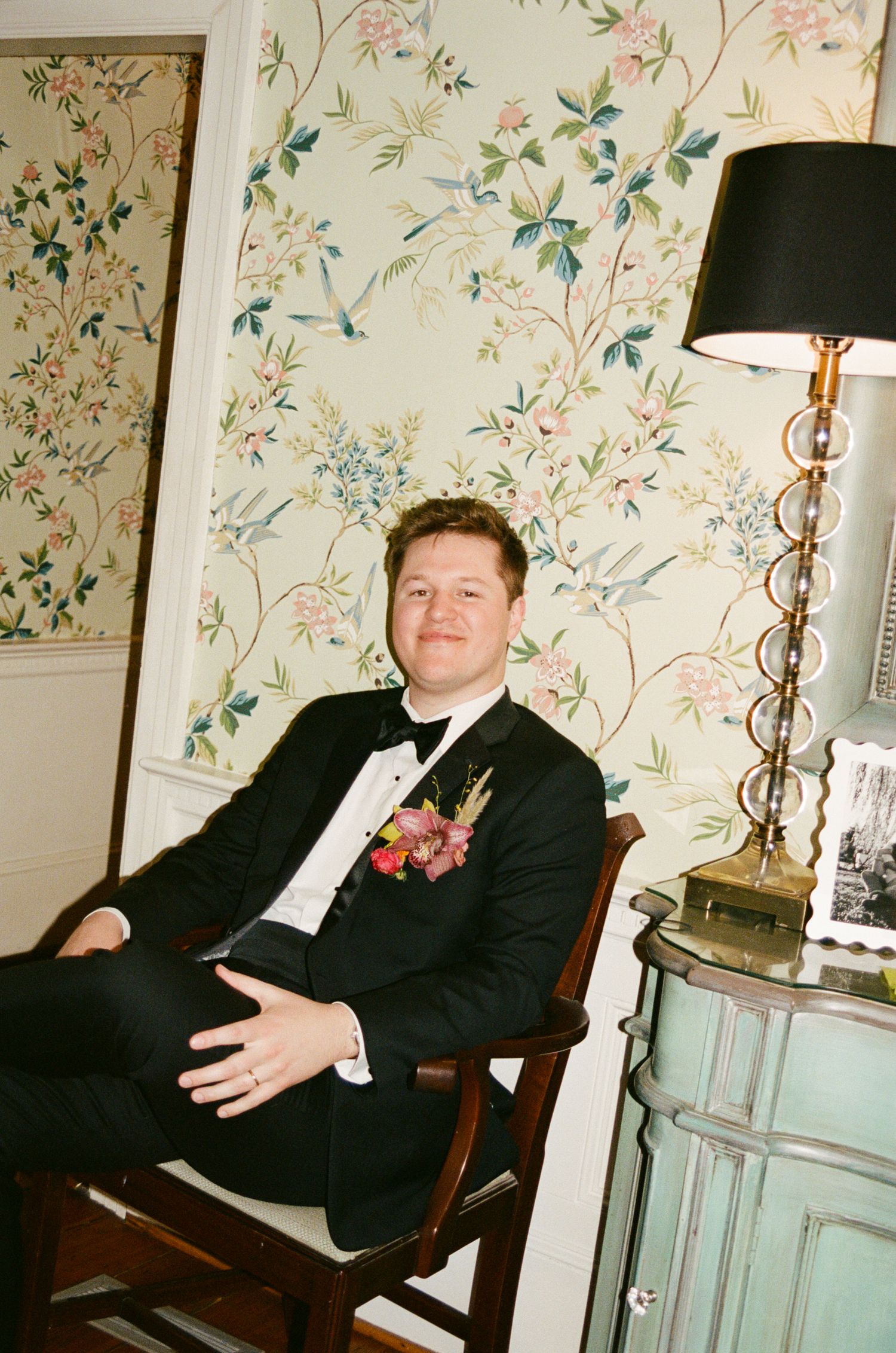 A groom in a black tuxedo with floral boutonniere sits in a wooden chair beside a lamp against floral wallpaper.