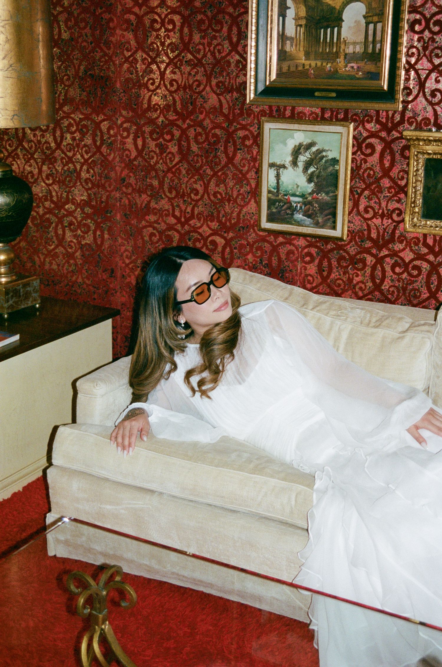 A person in a white dress lies asleep on a vintage red velvet couch in an ornate room with patterned wallpaper.