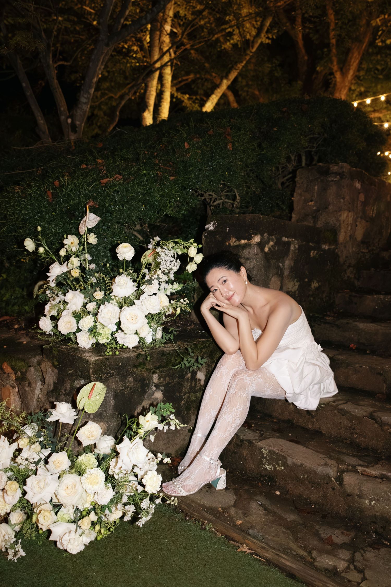 A romantic garden scene at night with stone steps adorned with lush white floral arrangements and string lights in the trees.