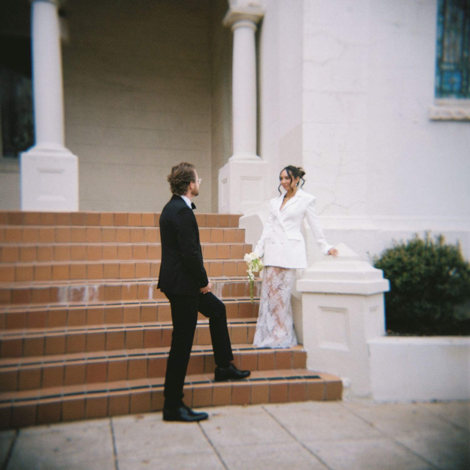 A bride in a white blazer dress holds a bouquet while facing a groom in a black suit on elegant building steps.