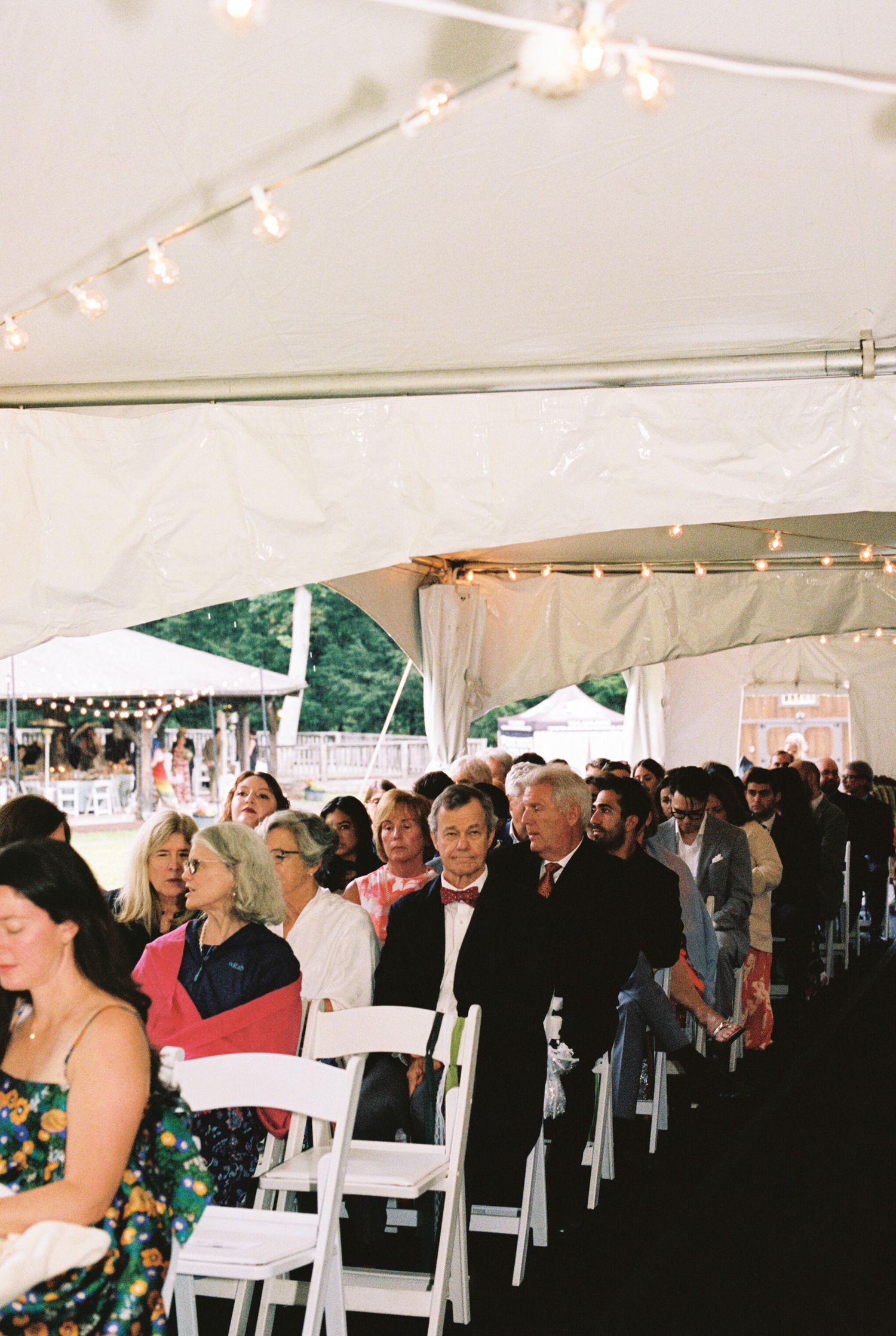 Wedding guests seated in white chairs under a tent with string lights and decorative lighting at an outdoor ceremony.