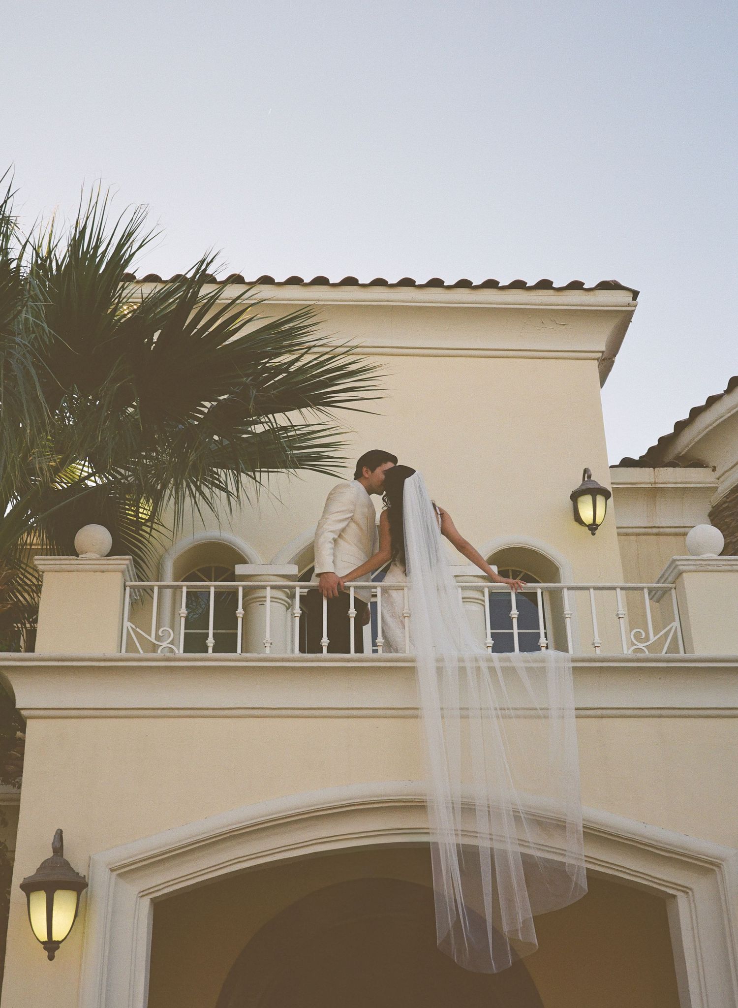 A couple shares a romantic moment on a Mediterranean-style balcony with palm trees and warm lighting at dusk.