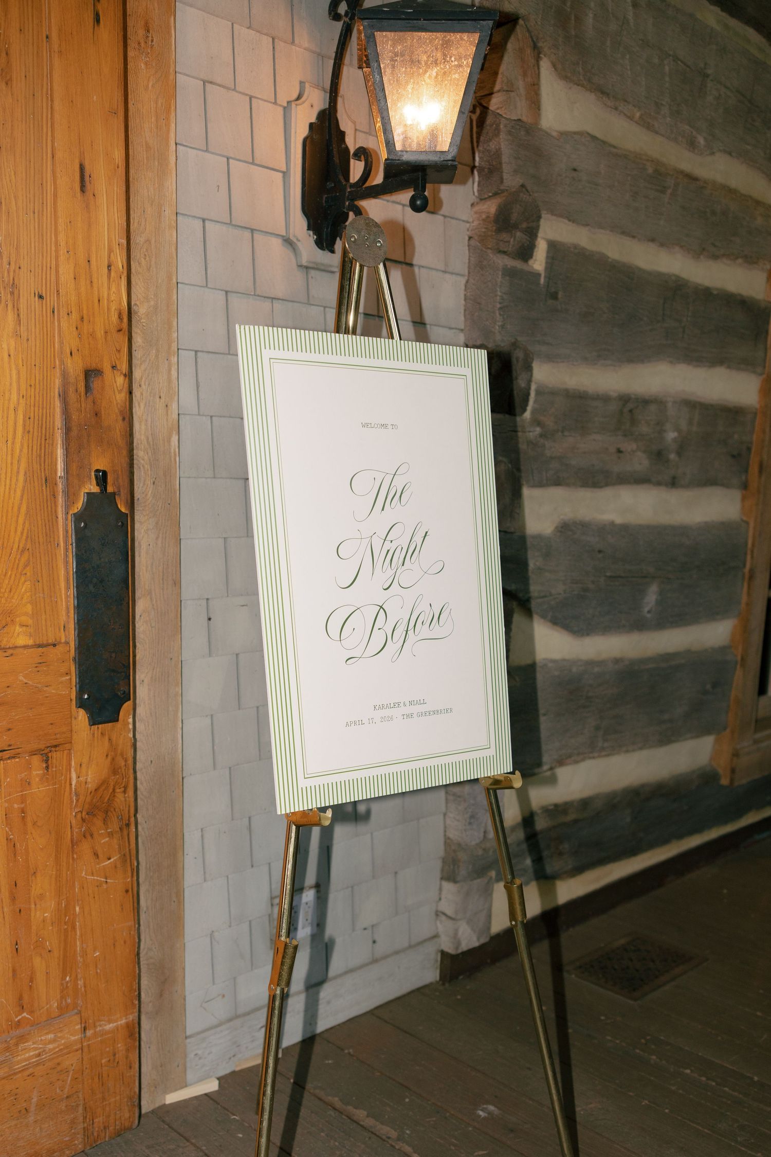 Elegant gold easel displaying a calligraphy wedding sign reading The Night Before against a rustic log cabin wall.