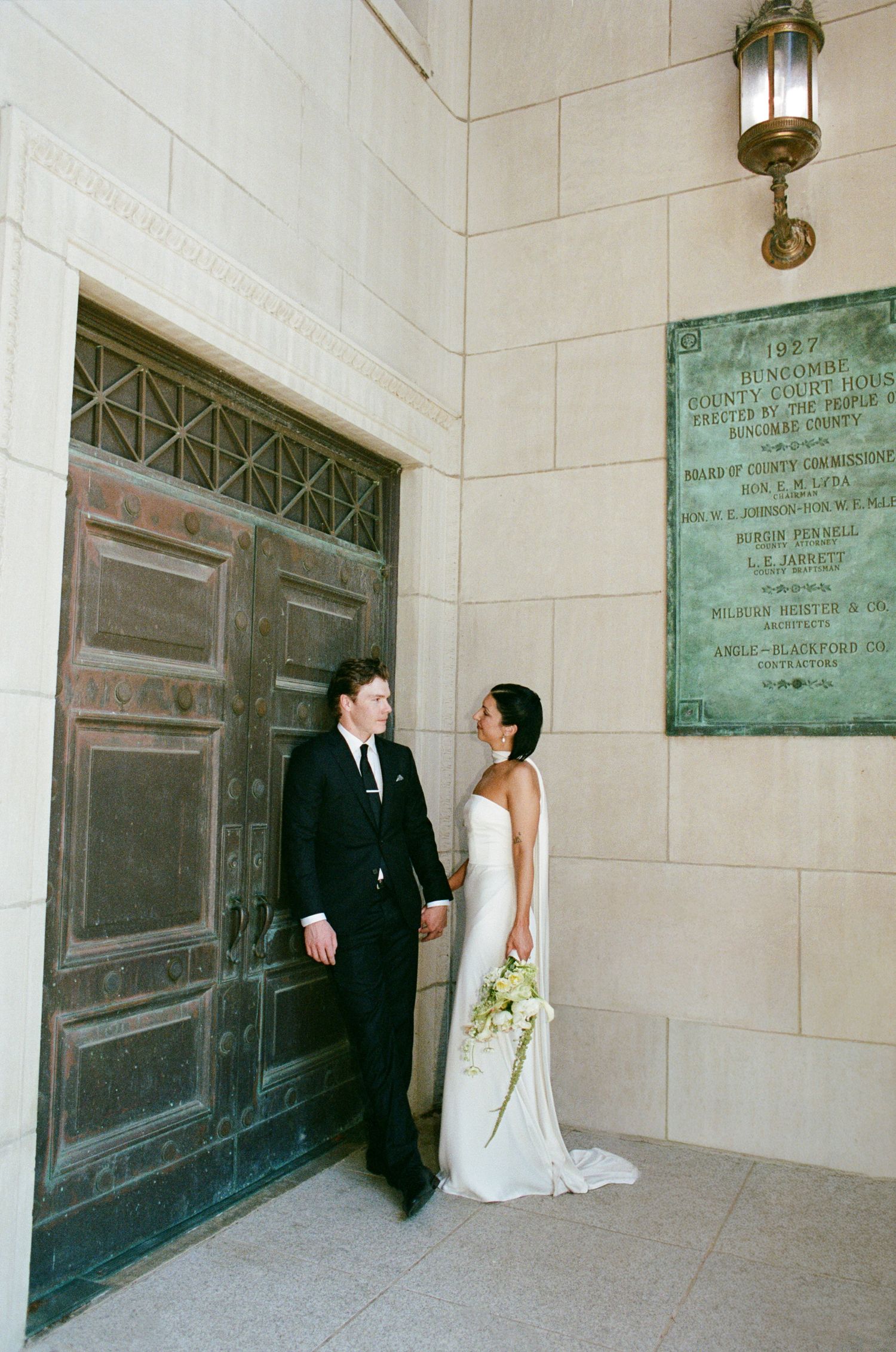 Bride in a strapless white gown holding a bouquet stands beside a groom in a black suit near an ornate bronze door and courthouse plaque.