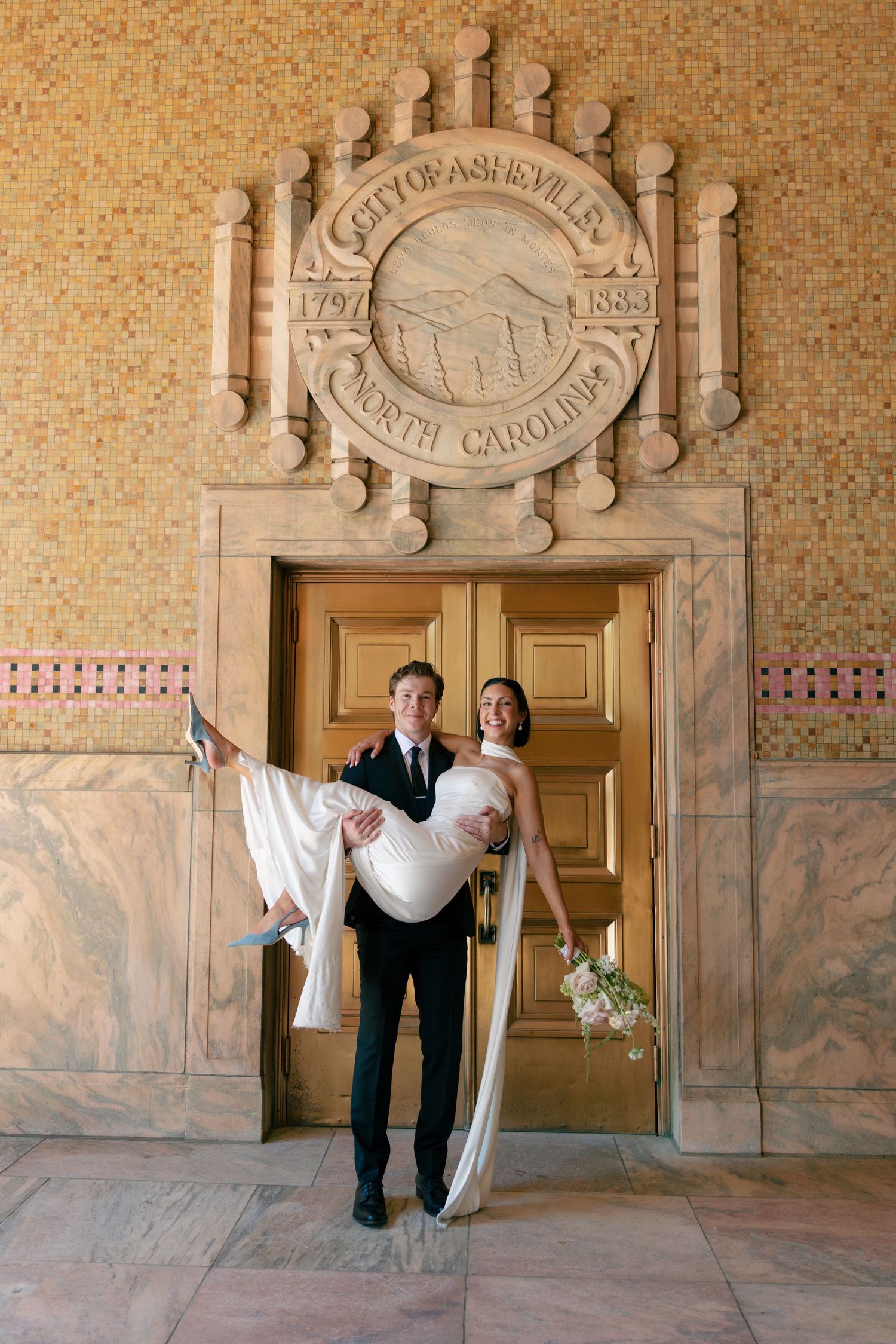 Groom in a dark suit carries a bride in a white gown and holds her bouquet beneath the City of Asheville, North Carolina seal at City Hall.