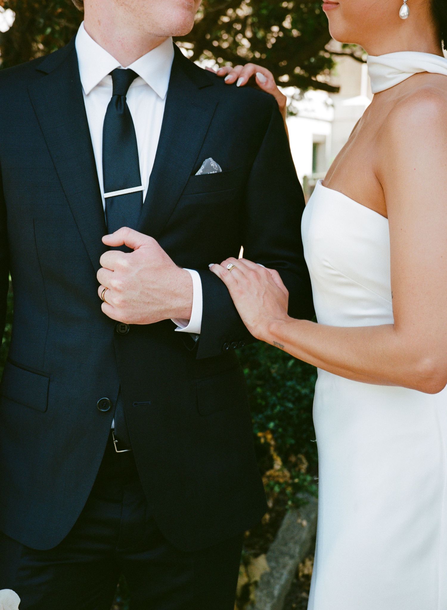 Bride in a white halter gown rests her hand on a groom in a navy suit, black tie and pocket square in a close wedding portrait.