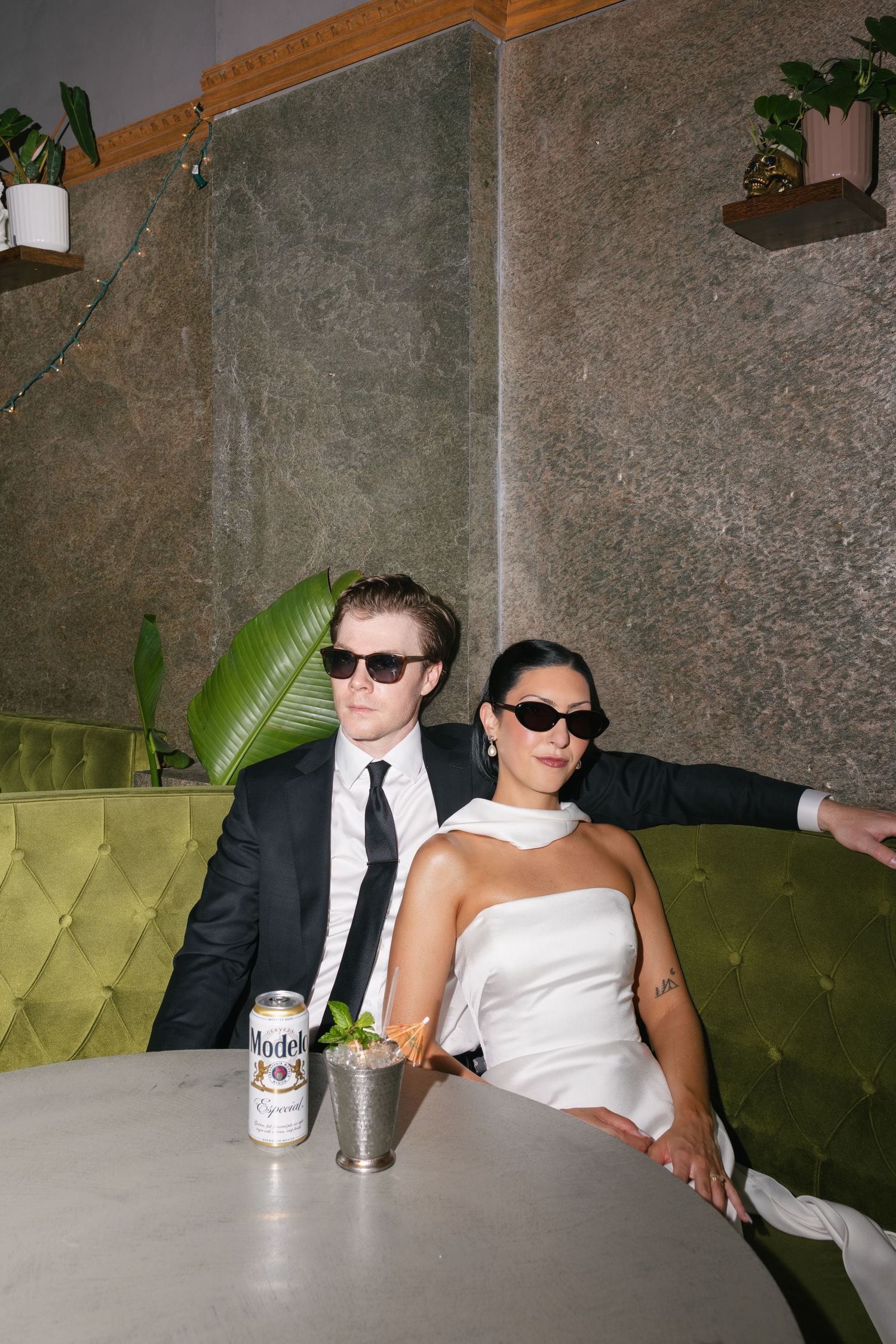 Couple in sunglasses and formal wedding attire sit on a green tufted velvet booth beside a marble table with a Modelo can and a mint julep.