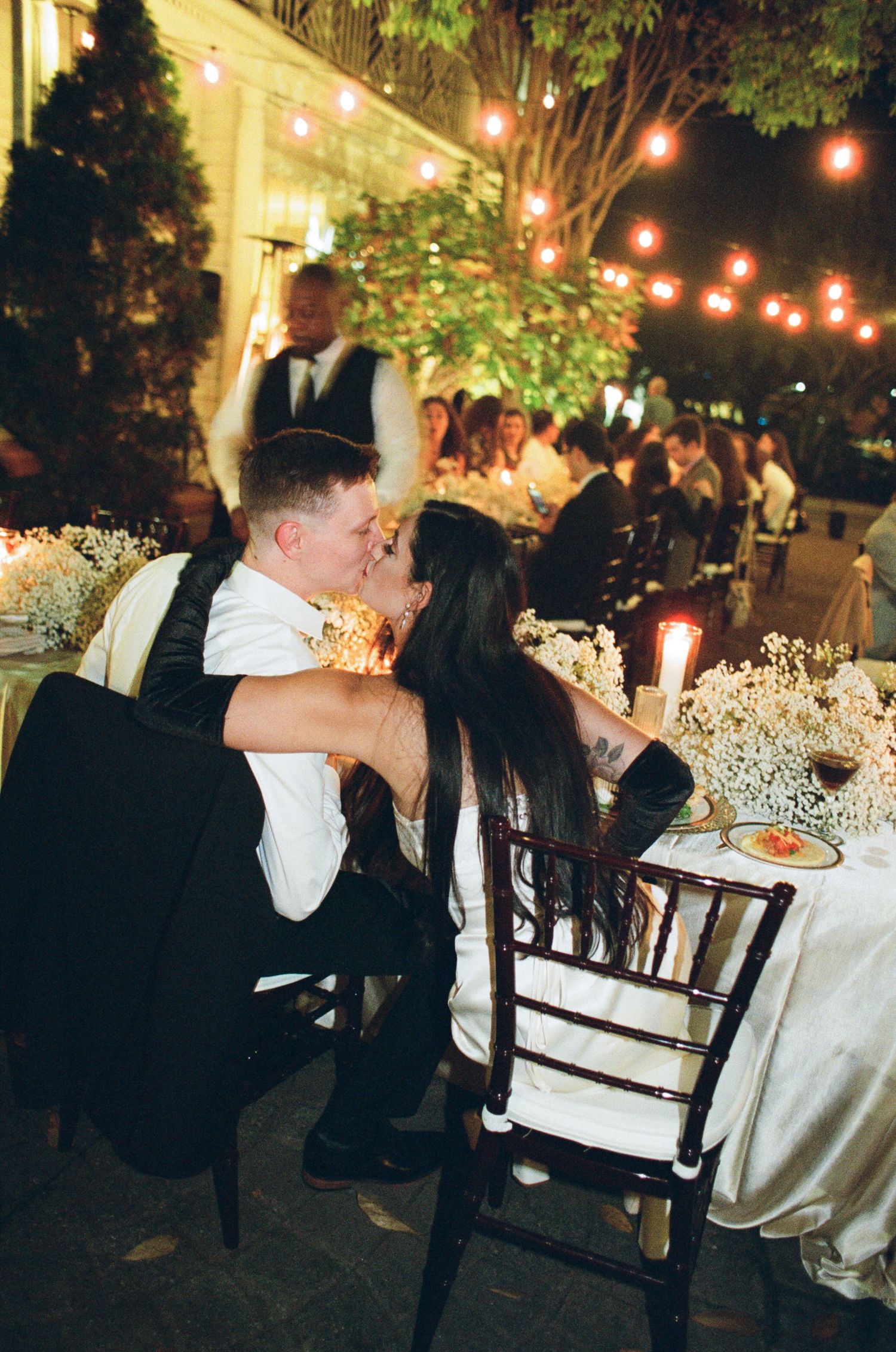 Couple shares a kiss at an outdoor evening wedding reception with string lights, candles, white floral centerpieces and guests at tables.