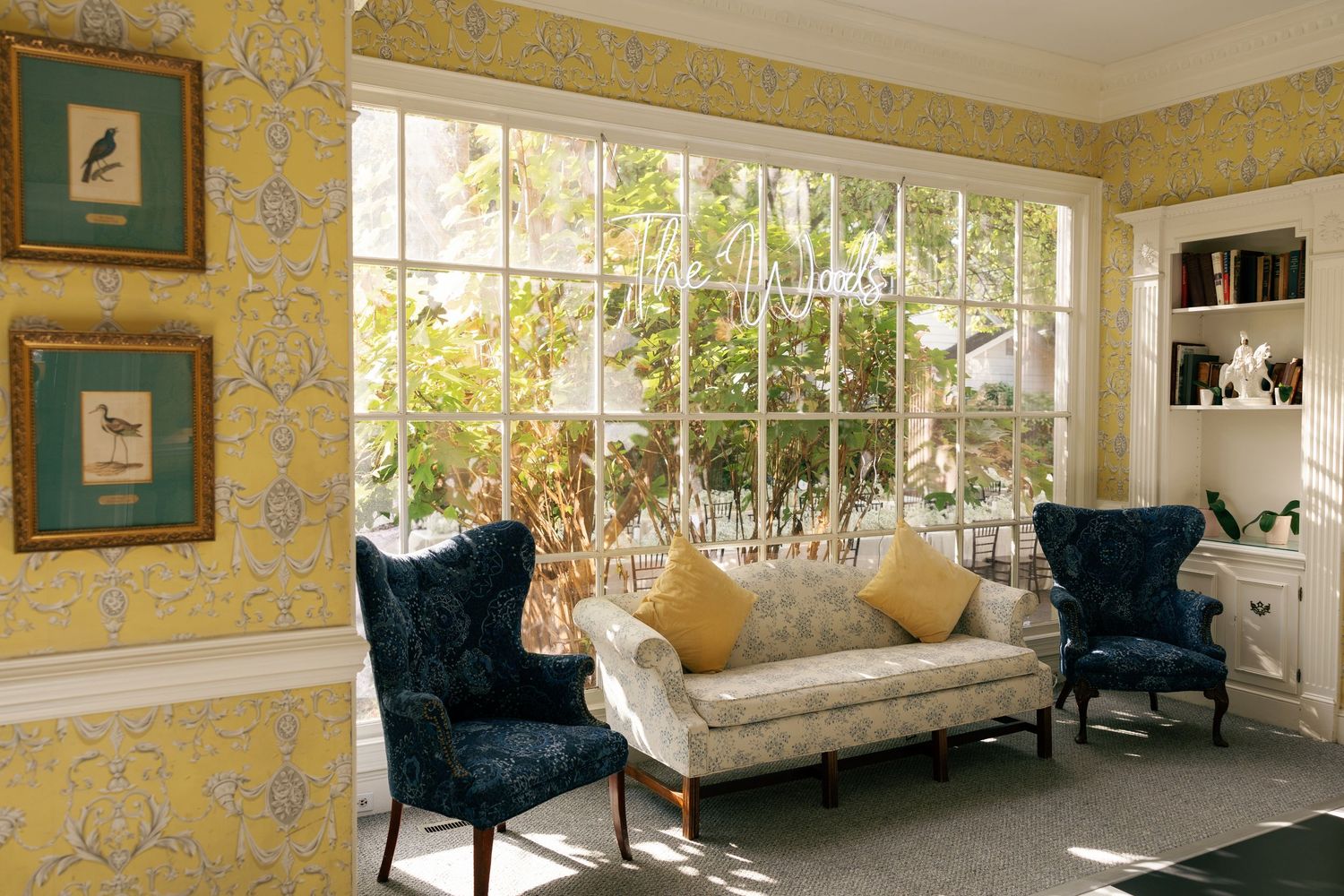Sunlit sitting room with yellow floral wallpaper, navy wingback chairs, a cream sofa with gold pillows, bird prints and large windows.