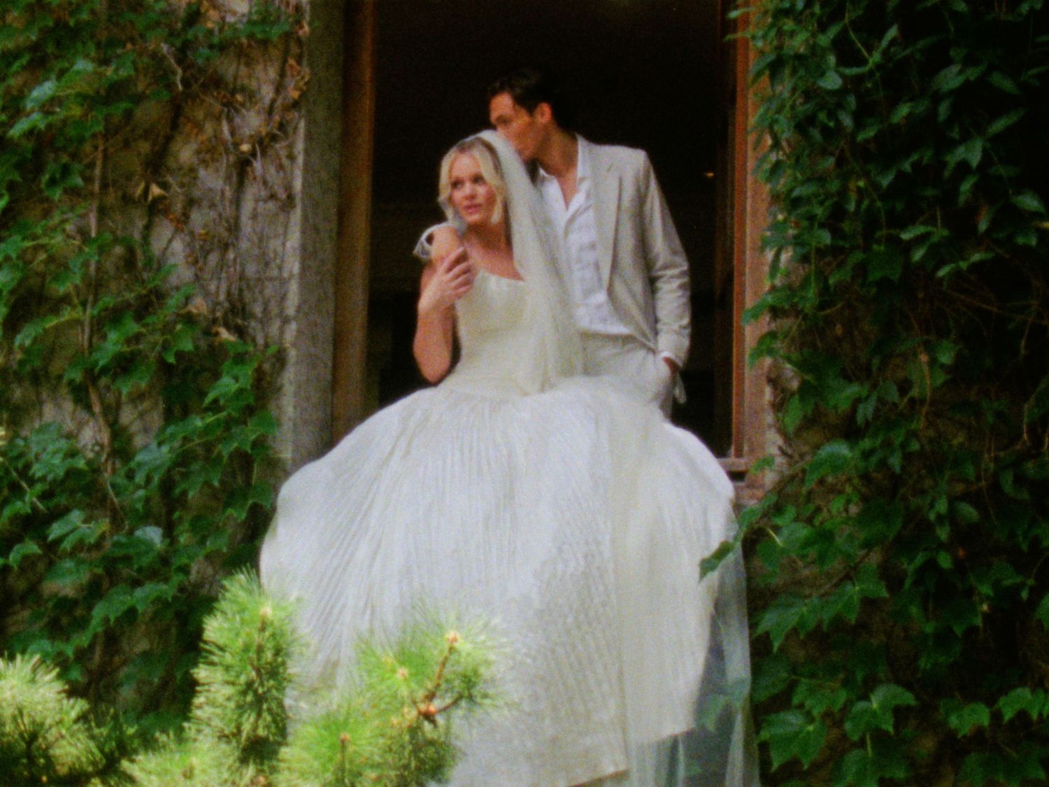 Bride in white ball gown and groom in gray suit stand in doorway surrounded by lush greenery on wedding day.