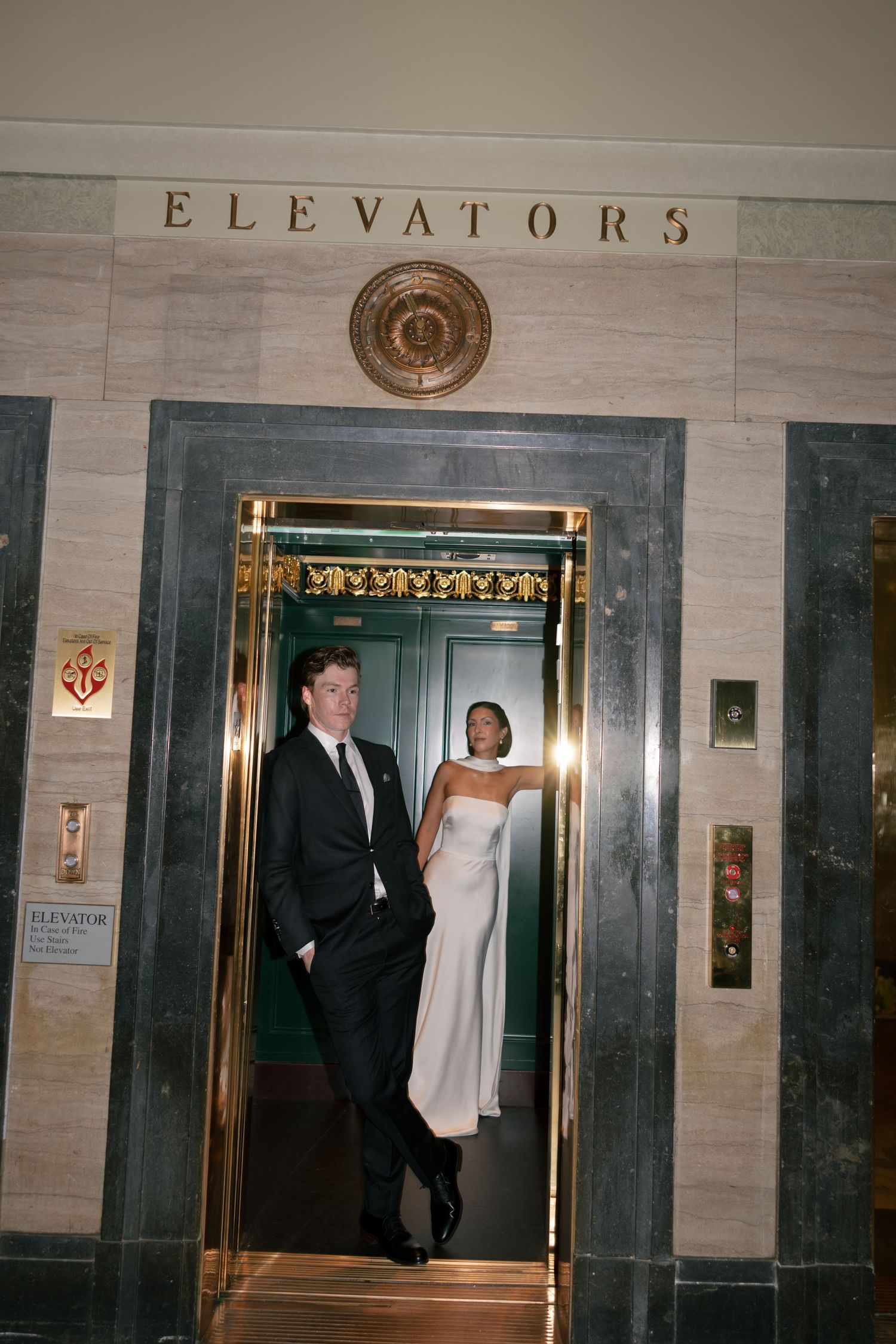 A man in a dark suit and a woman in a white gown pose in an open brass elevator beneath an ornate marble elevators sign.