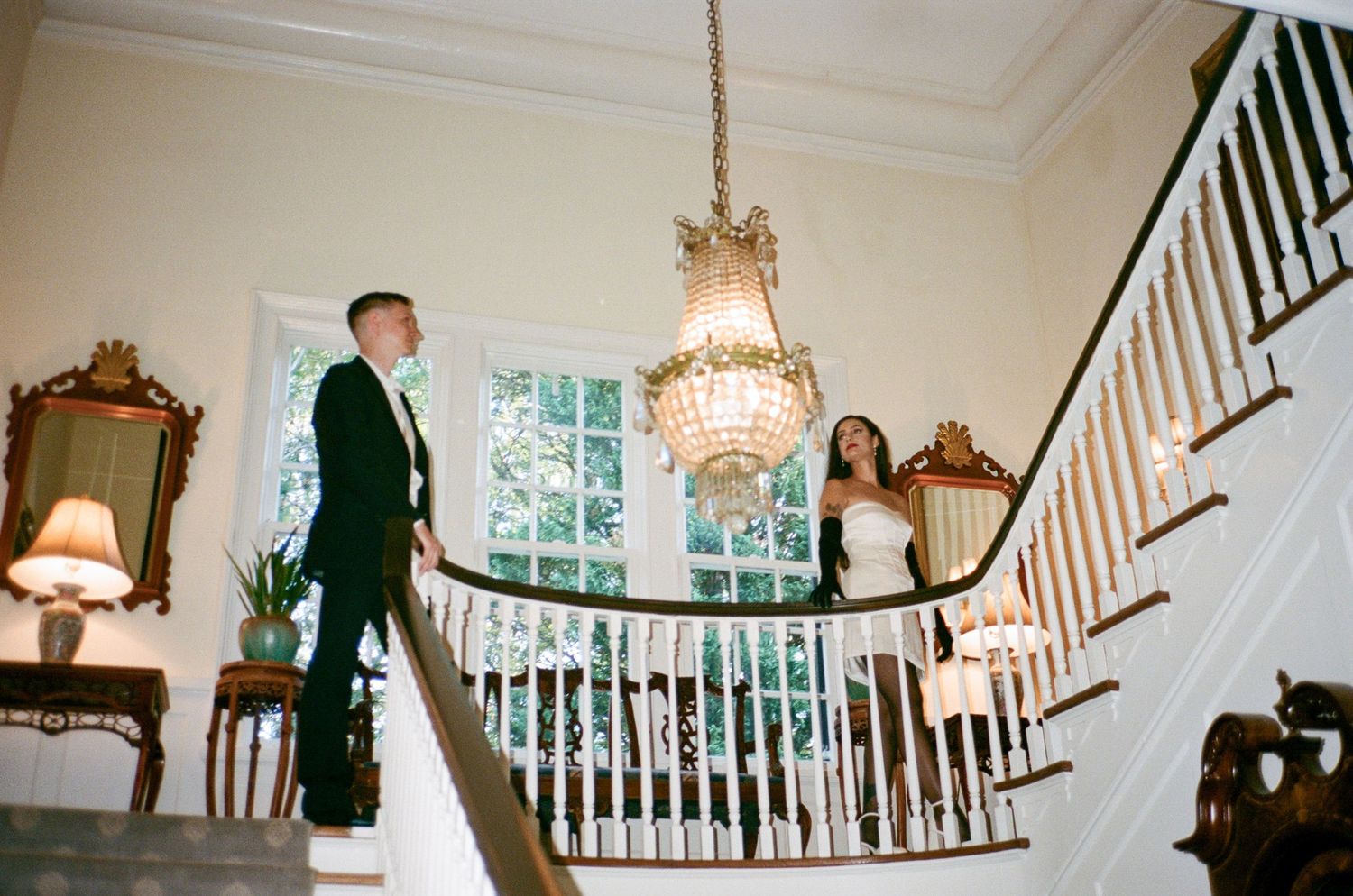 Bride and groom pose on an elegant curved staircase beneath a crystal chandelier in a grand historic venue with ornate mirrors.