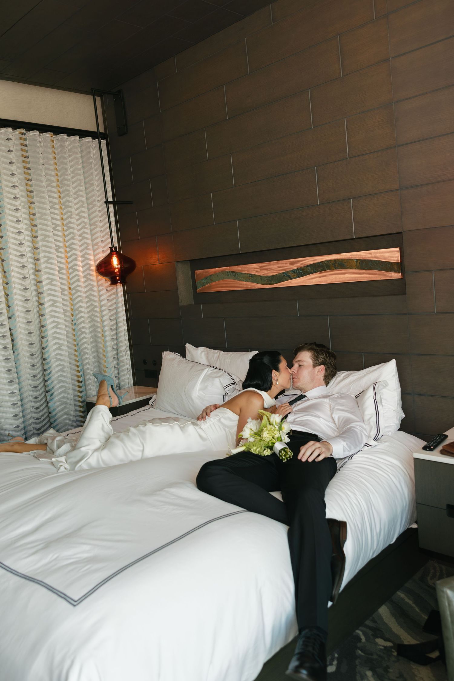 Newlywed couple in wedding attire kissing on a white hotel bed with a bridal bouquet in a modern room with dark wood paneling.