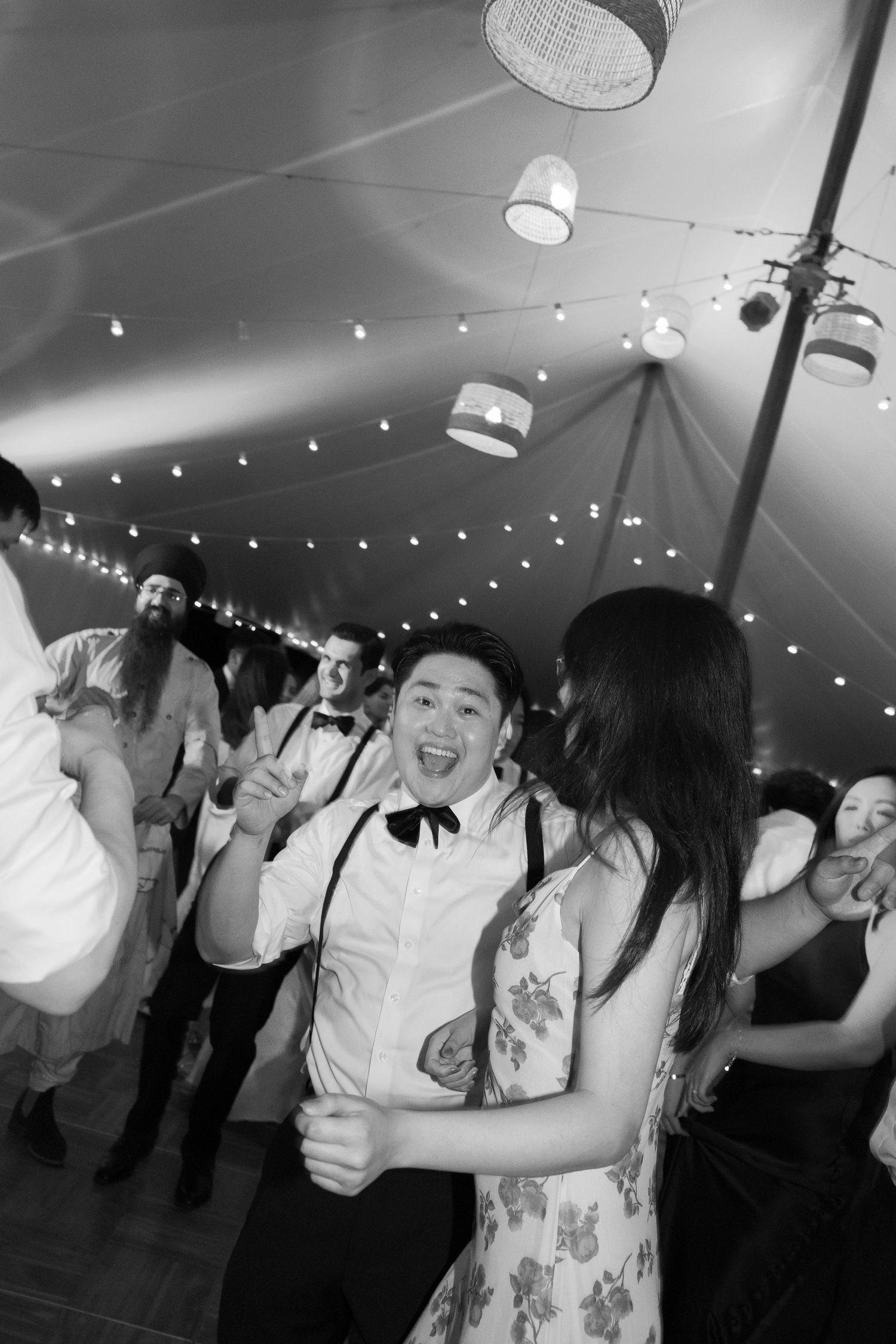 People dancing and celebrating under a tent with string lights and drum shade pendant lamps at a festive event.