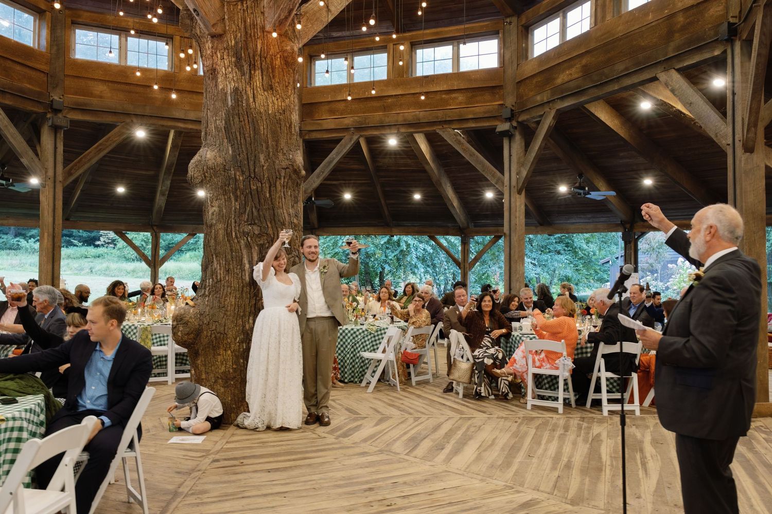 A couple dances their first dance under a rustic wooden pavilion while guests watch from white folding chairs.