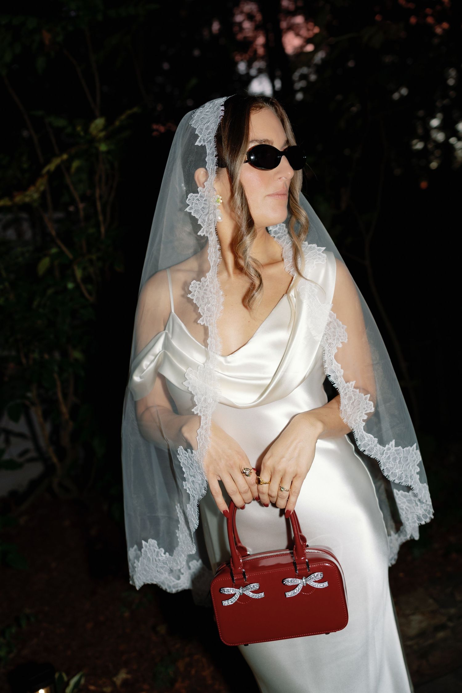 Wedding dress detail shot with red handbag showing delicate lace veil and white satin fabric against dark background.