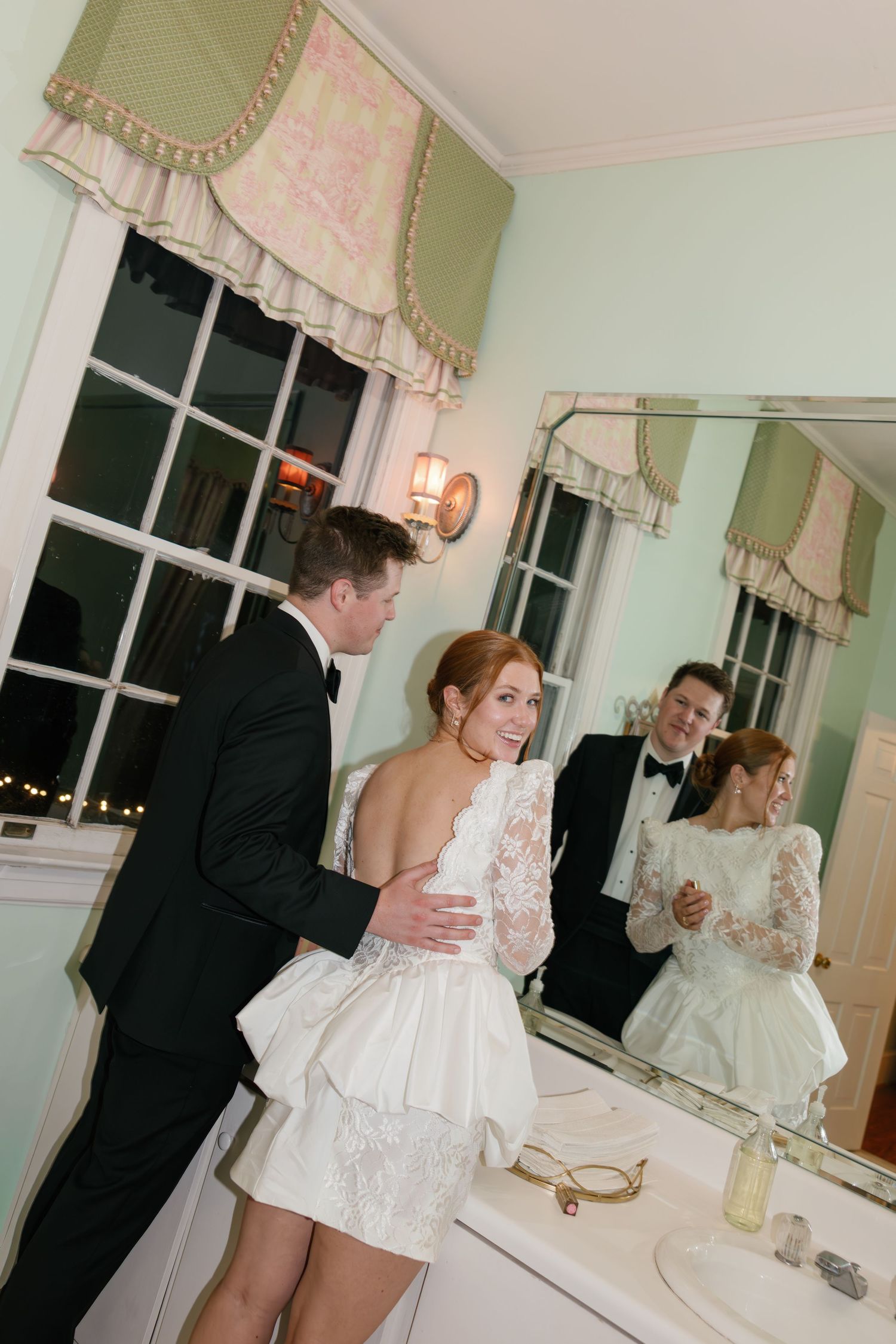 Bride and groom share a romantic moment in an elegant bathroom, their reflection visible in the mirror.