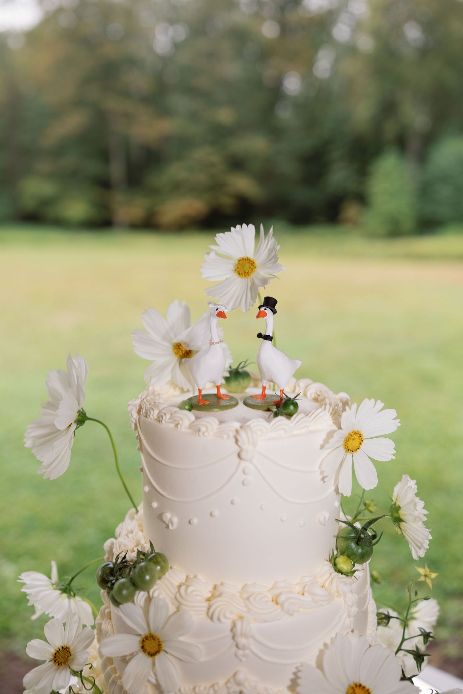 White two-tier wedding cake decorated with fresh daisies and delicate piping details, topped with miniature figures outdoors.