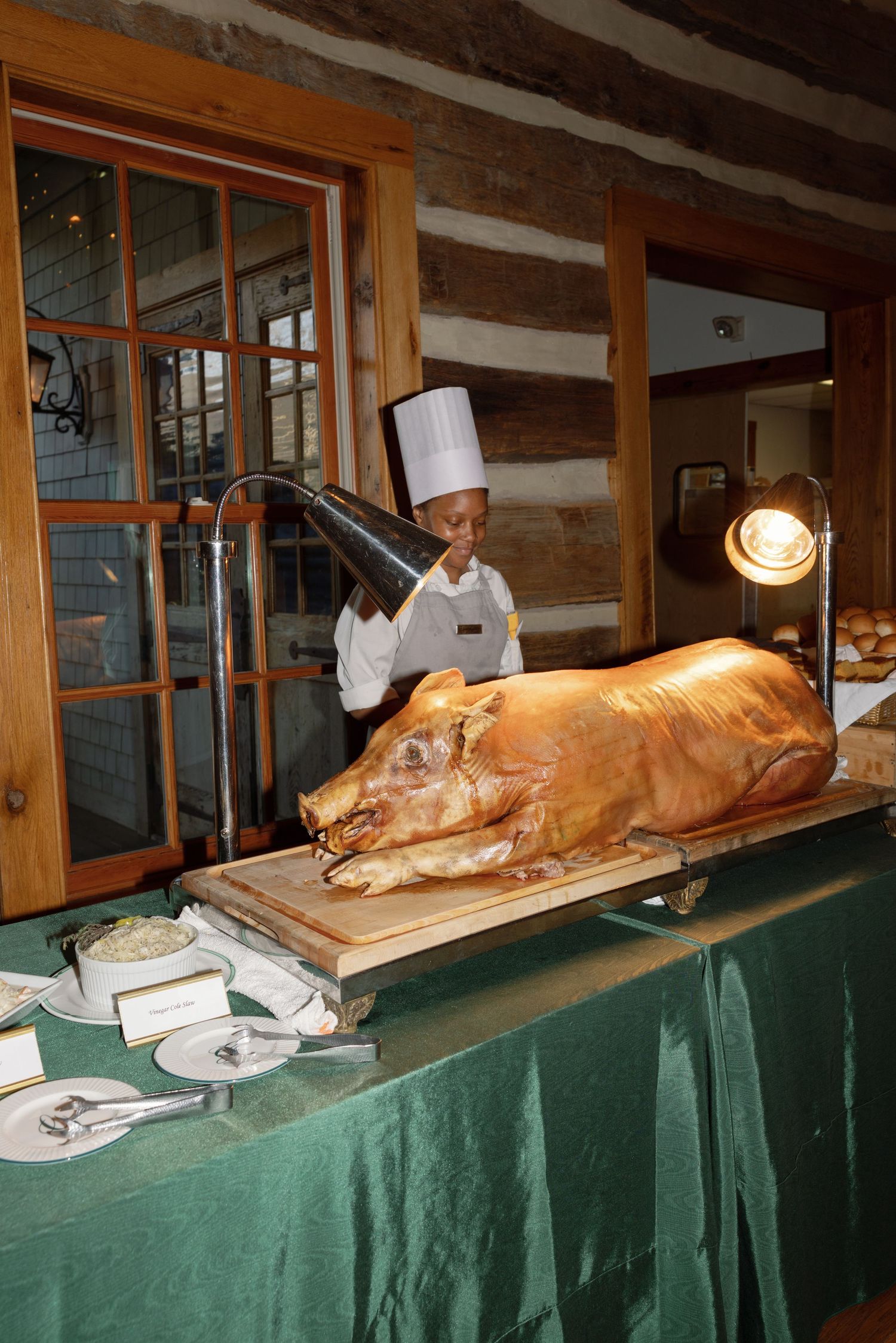 Chef in white hat stands behind a whole roasted pig on a green draped buffet table in a rustic restaurant.
